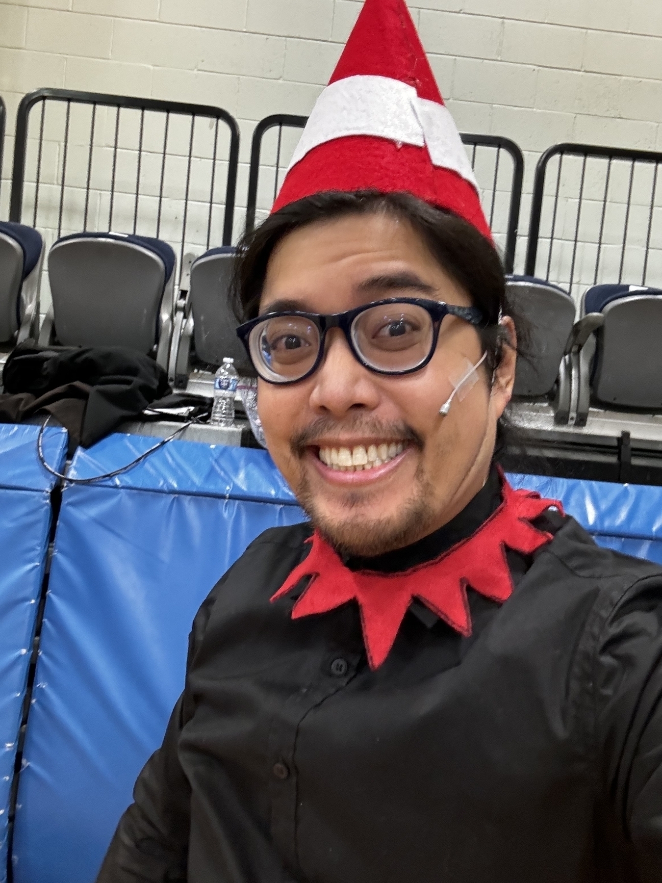 Miguel a masc Filipino man wearing a pointy red elf hat and black shirt and smiling