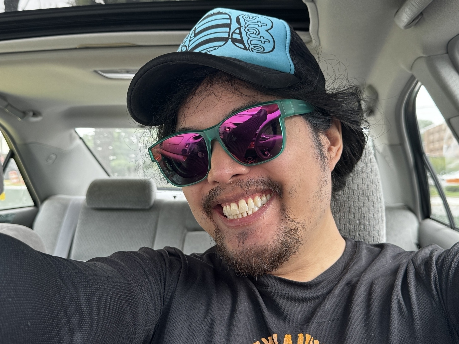 Miguel a masc Filipino man is wearing a black shirt inside a car. He is wearing purple lens sunglasses with a green frame. And a blue trucker hat. He is smiling with teeth. His hair is in a ponytail. It is day time. You set him from the chest up. 