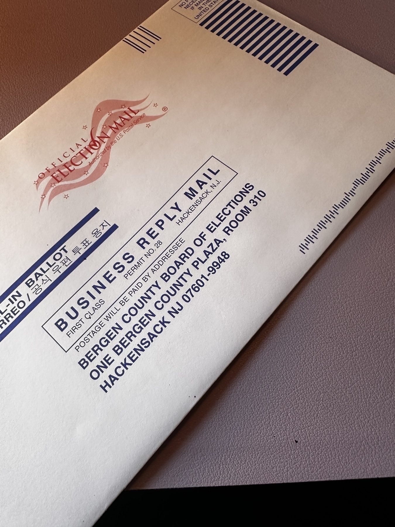 Envelope sat on a Desk. Desk mat is lavender. Envelope is diagonal to frame. It is a white envelope with blue font. It is indoors. &10;&10;BALLOT&10;BUSINESS REPLY MAIL&10;FIRST CLASS&10;PERMIT NO. 28&10;HACKENSACK, N.J.&10;POSTAGE WILL BE PAID BY ADDRESSEE&10;BERGEN COUNTY BOARD OF ELECTIONS&10;ONE BERGEN COUNTY PLAZA, ROOM 310&10;HACKENSACK NJ 07601-9948&10;