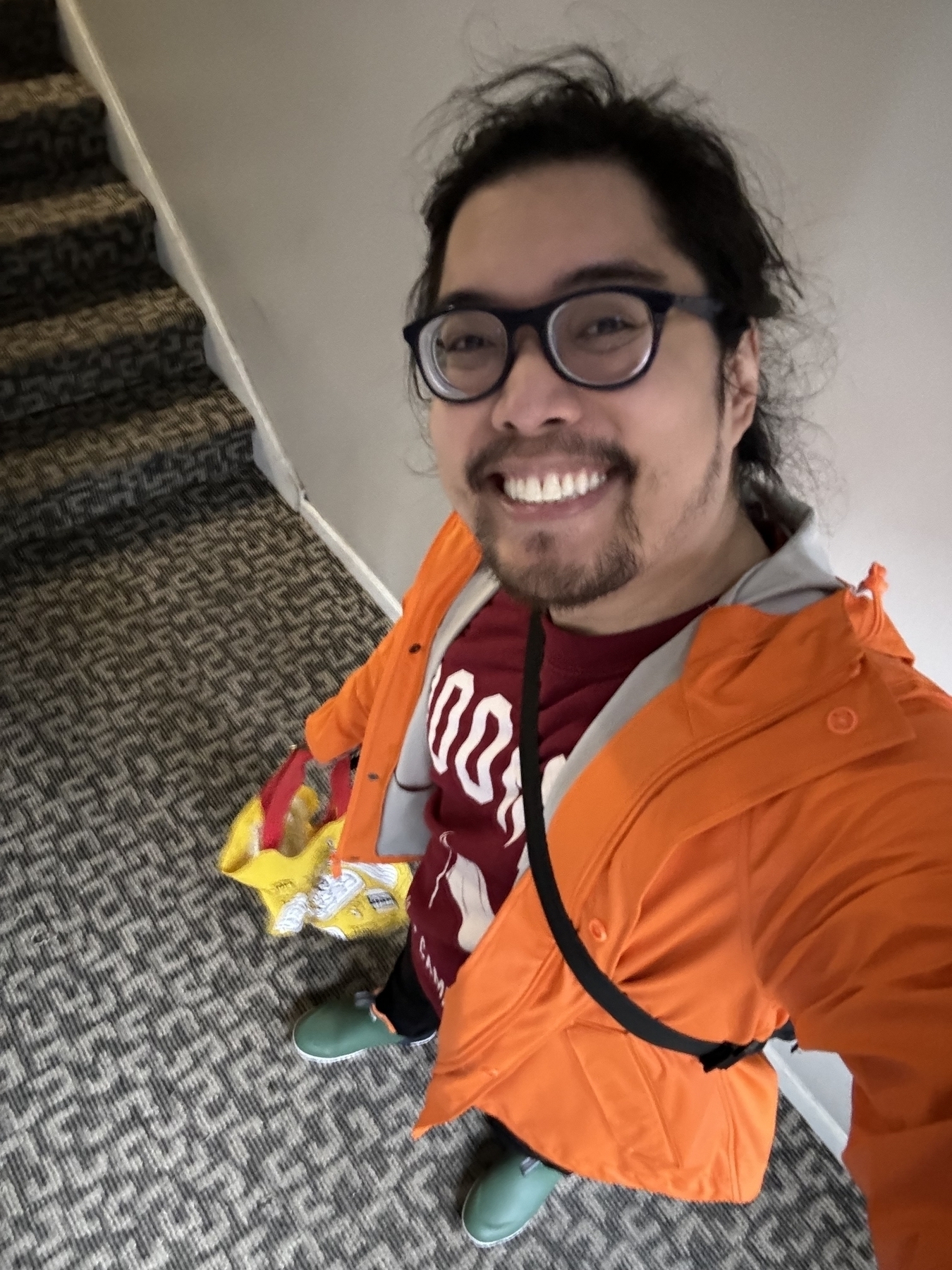 Man taking a selfie in a corridor, wearing glasses, a red shirt, orange jacket, and holding a yellow bag, with a staircase in the background.