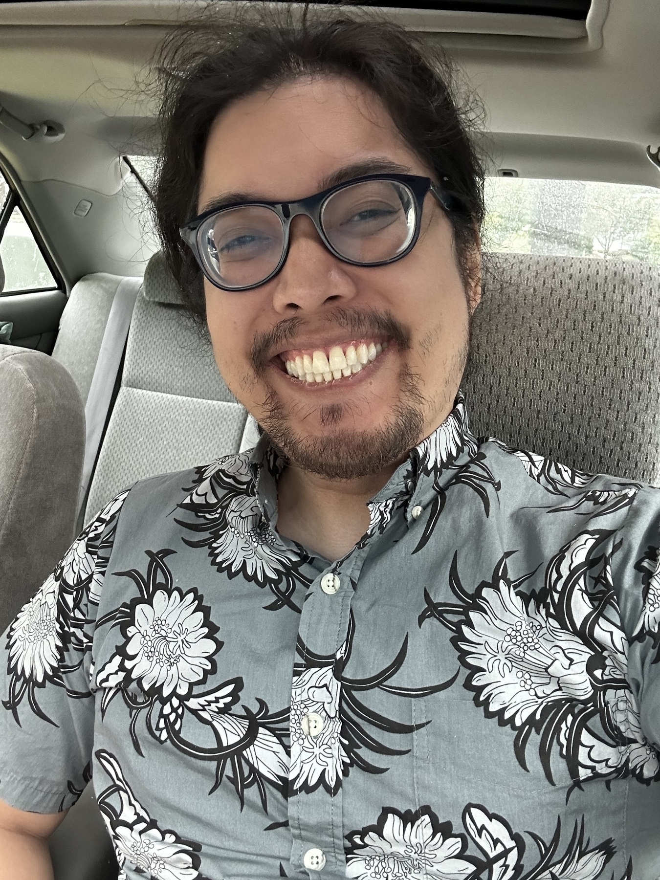 Selfie in a car by Miguel a Filipino man with glasses and black hair in a ponytail. He is smiling with teeth looking at camera. He is wearing a Hawaiian shirt that is grey with white flowers.