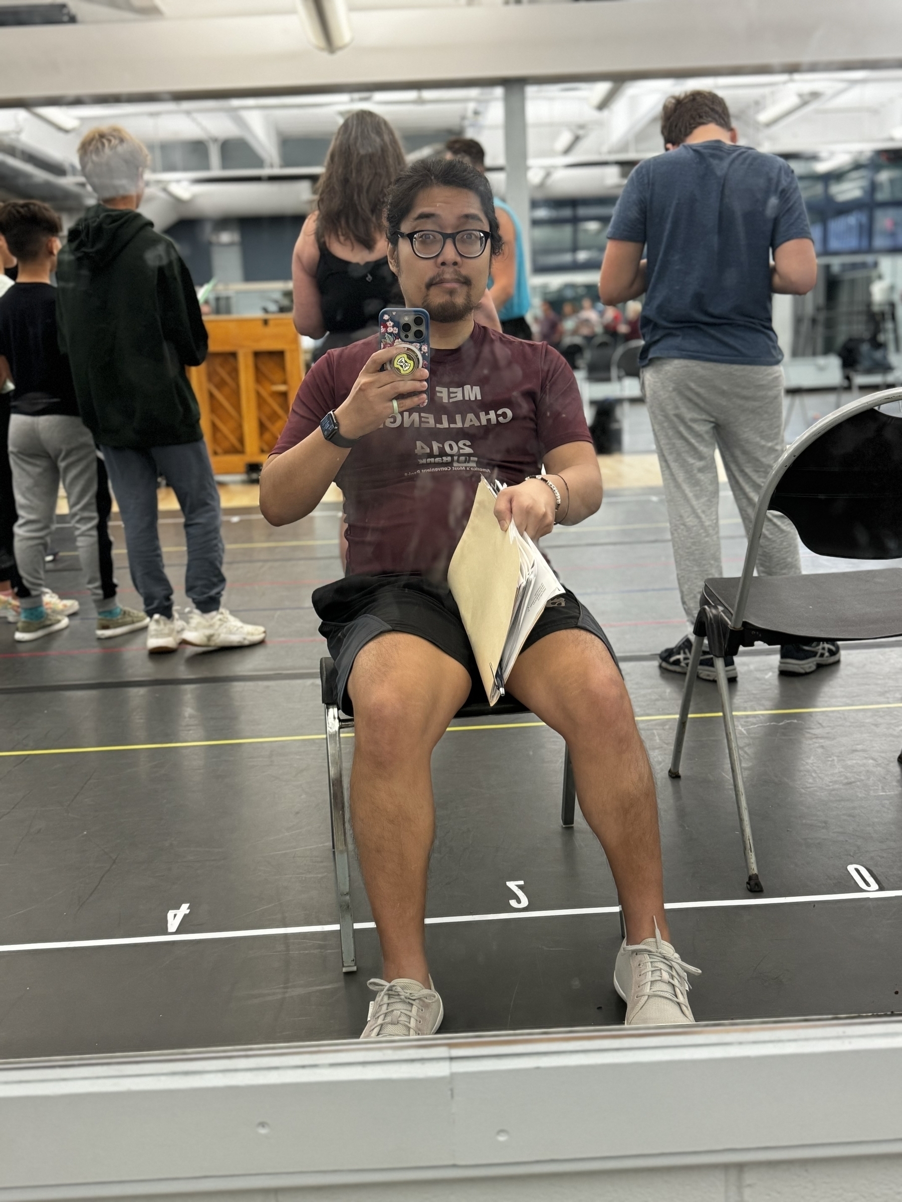 Miguel a masc Filipino man is sitting on a chair taking a photo of himself with a phone in a floor to ceiling mirror. He is wearing black shorts, glasses and a purple shirt. Behind him are his cast mates. His hair is in a pony tail. He is holding a Manila folder with his script. 