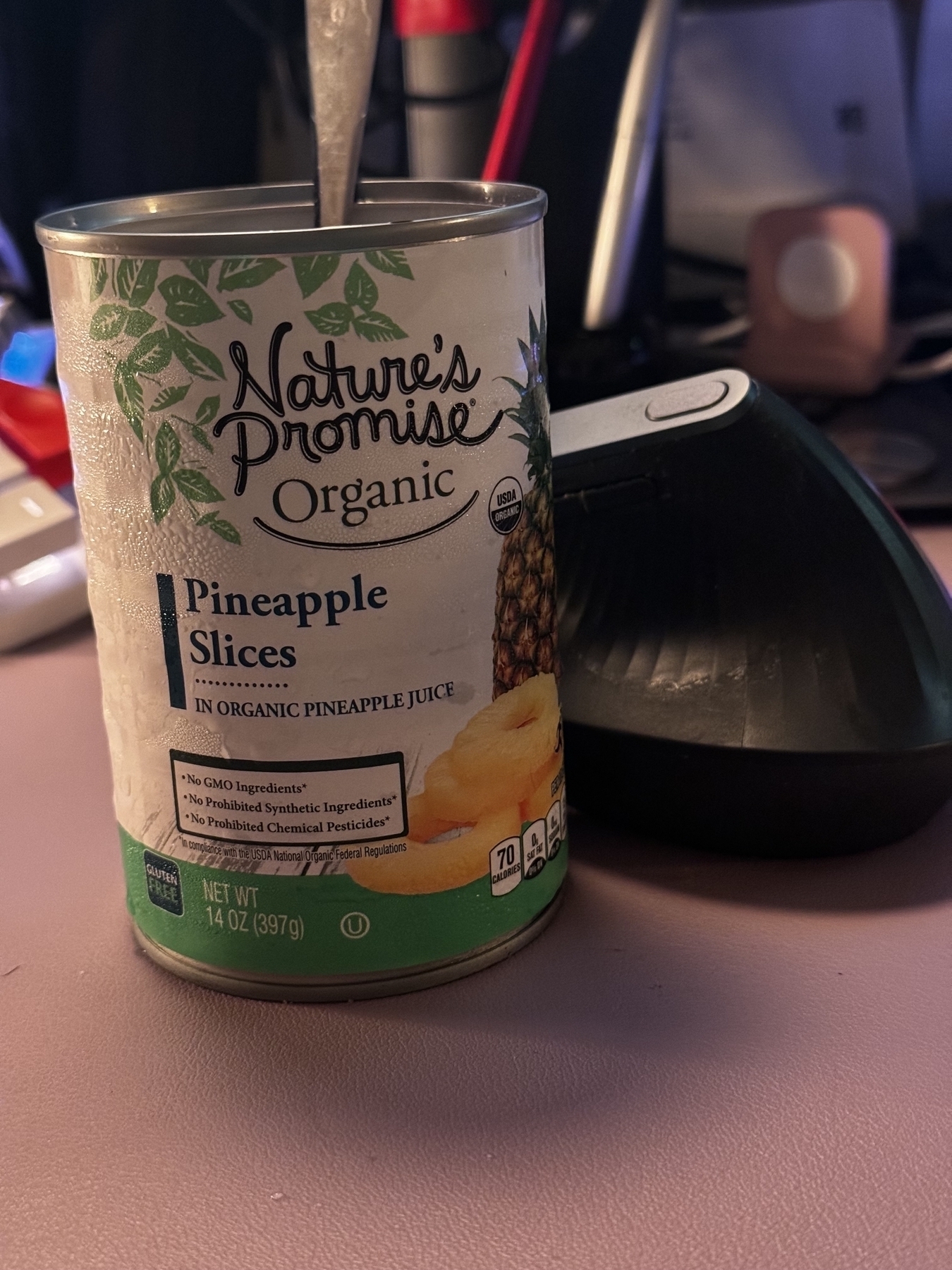 A can of organic natures promise store brand sliced pineapples sat on a lavender desk mat. A grey ergonomic mouse is sat next to it. A utensil is sticking out of the can. 