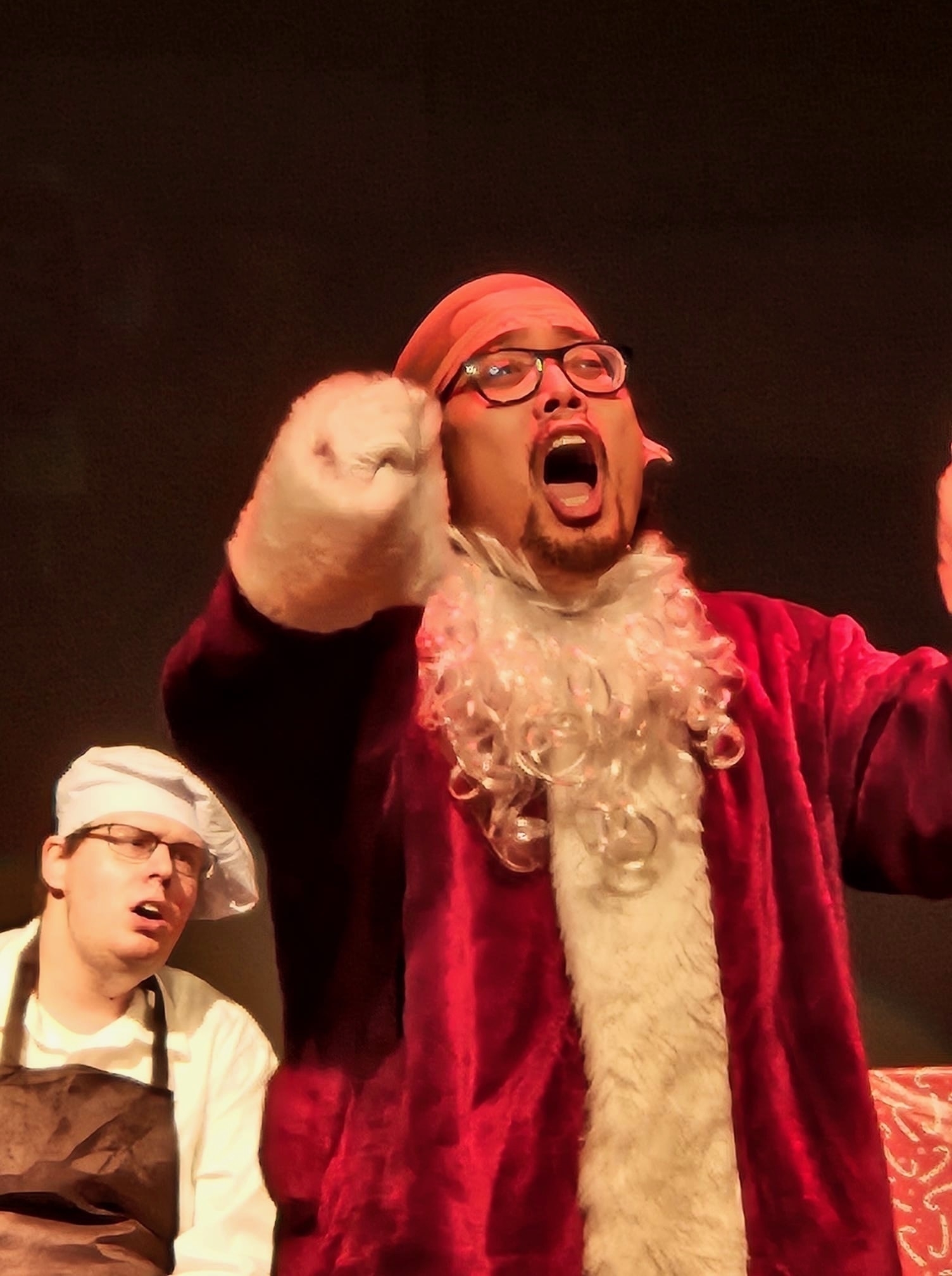 Two actors on stage, one in the foreground wearing a red costume with a white beard and glasses, appearing surprised or animated; the other in the background wearing a white chef hat and apron with a quizzical expression.
