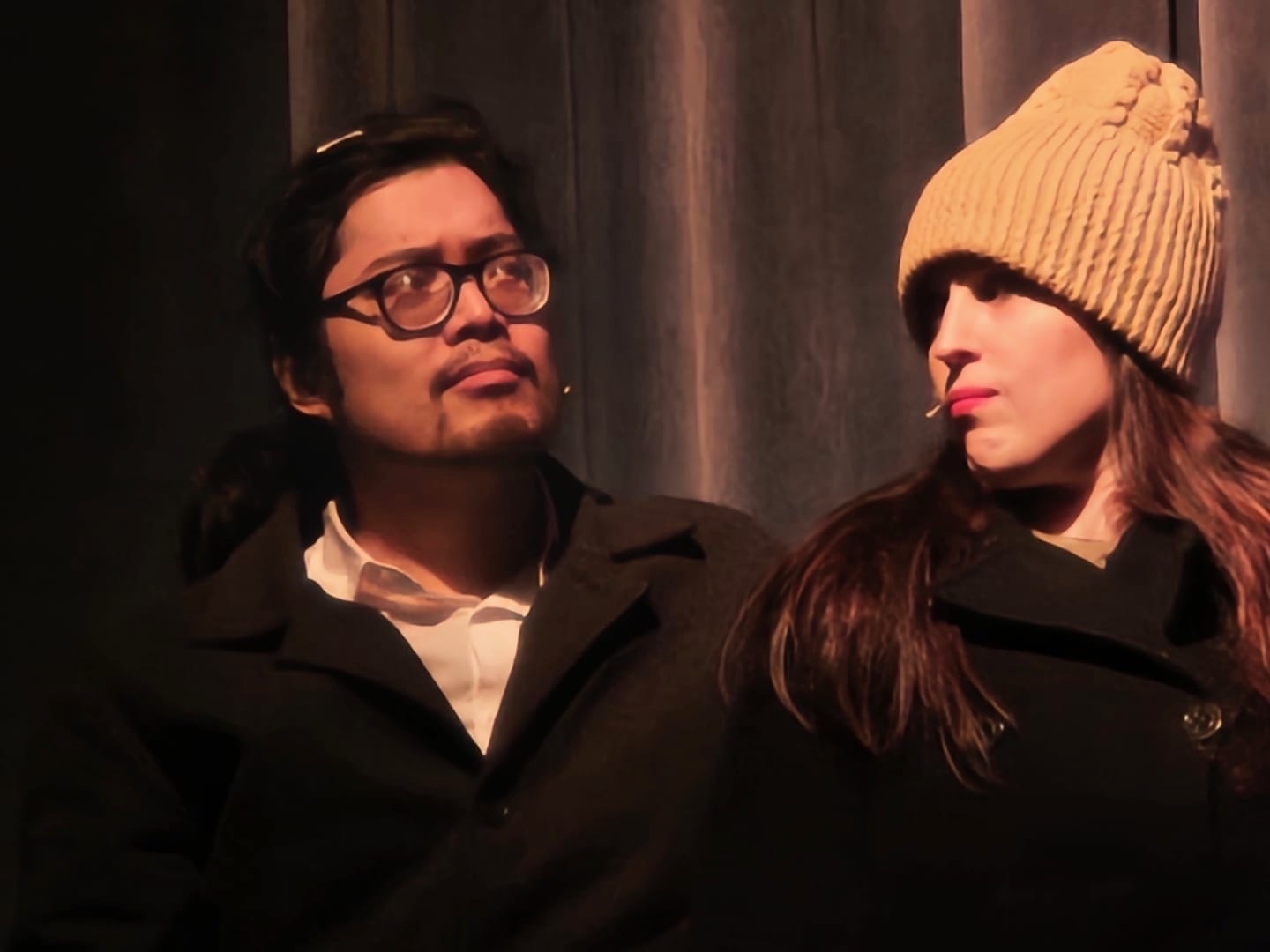 A man and a woman wearing winter attire, with the man looking up to the side and the woman looking away with a toothpick in her mouth. They appear to be standing indoors, with a dimly lit, curtain-like background.
