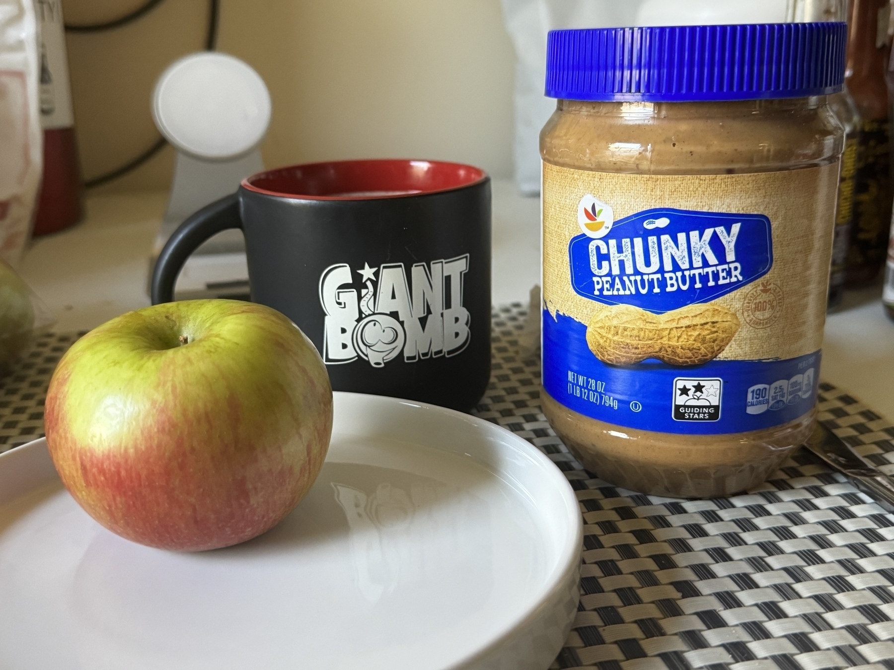 Three items. Honeycrisp apple on left on white platter. Giant bomb branded black mug with hazelnut coffee. On right store brand chunk peanut butter on a table indoors. 