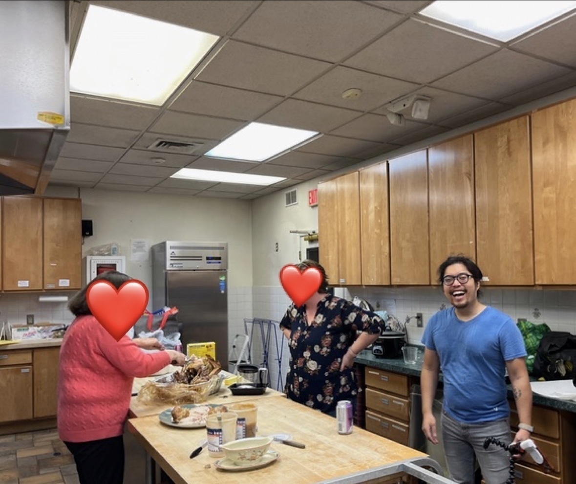 Three people in a kitchen. Miguel a Filipino man is to the right of frame. Wearing a blue shirt. Smiling big, mouth wide open. Holding a tripod. 