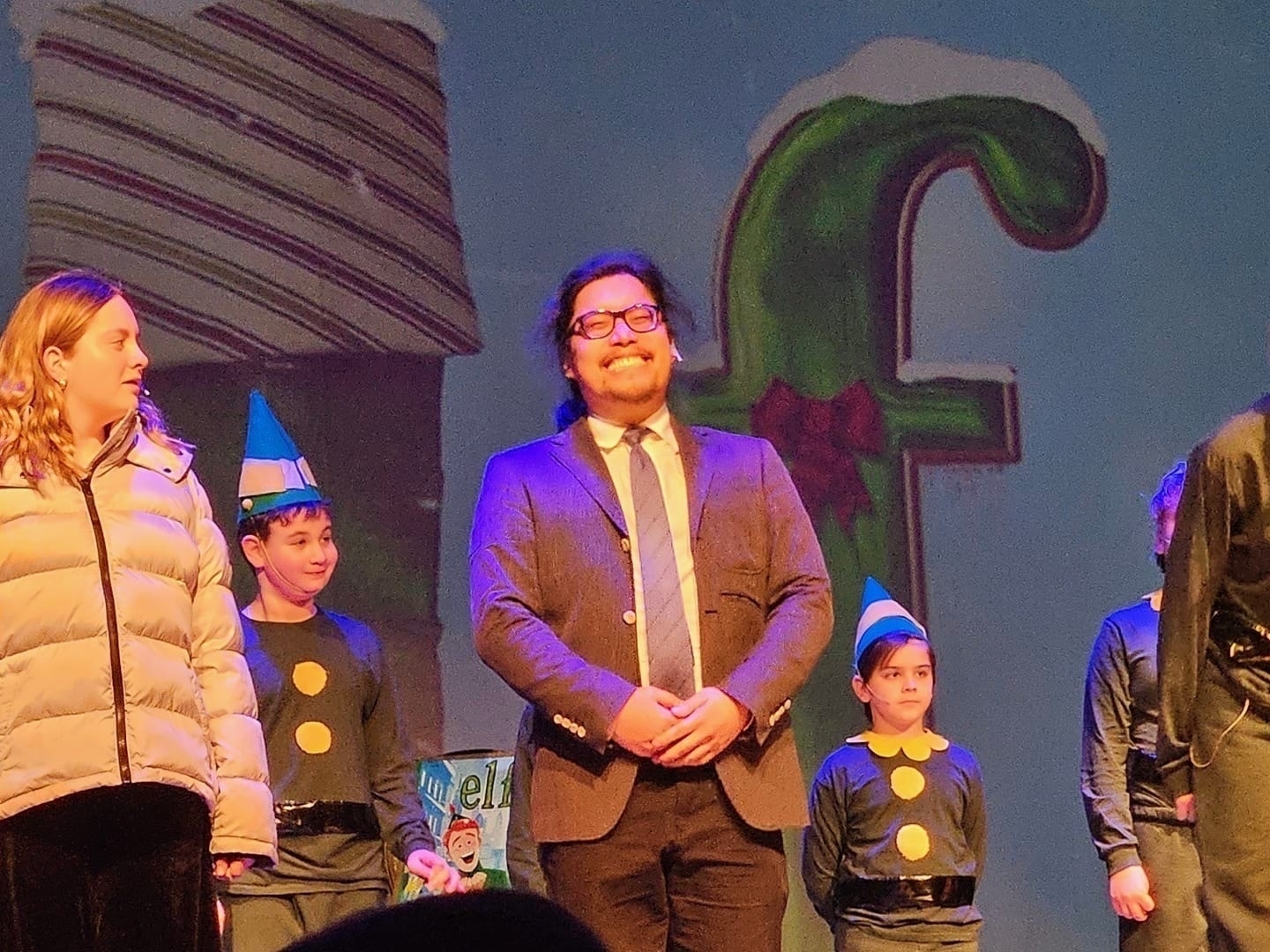 Group of people on stage with a man in a suit smiling at the center, flanked by individuals in elf costumes.