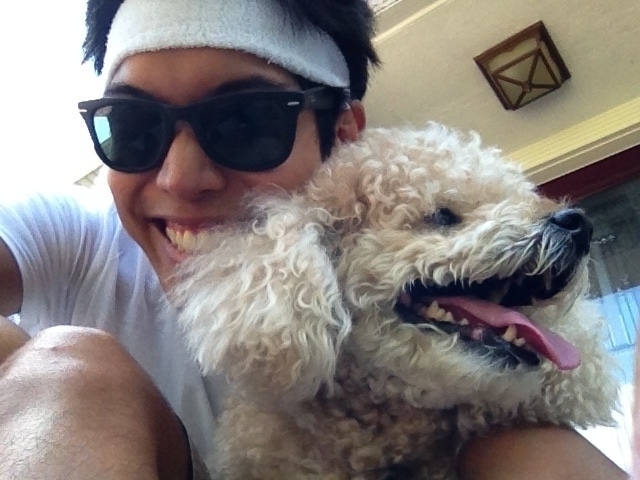 Selfie of Miguel a masc Filipino man smiling at camera hugging a light brown poodle. Poodle is looking to the right with his tongue out. Miguel is wearing black sunglasses and a white sweat head band. They are outside. It is daytime. 