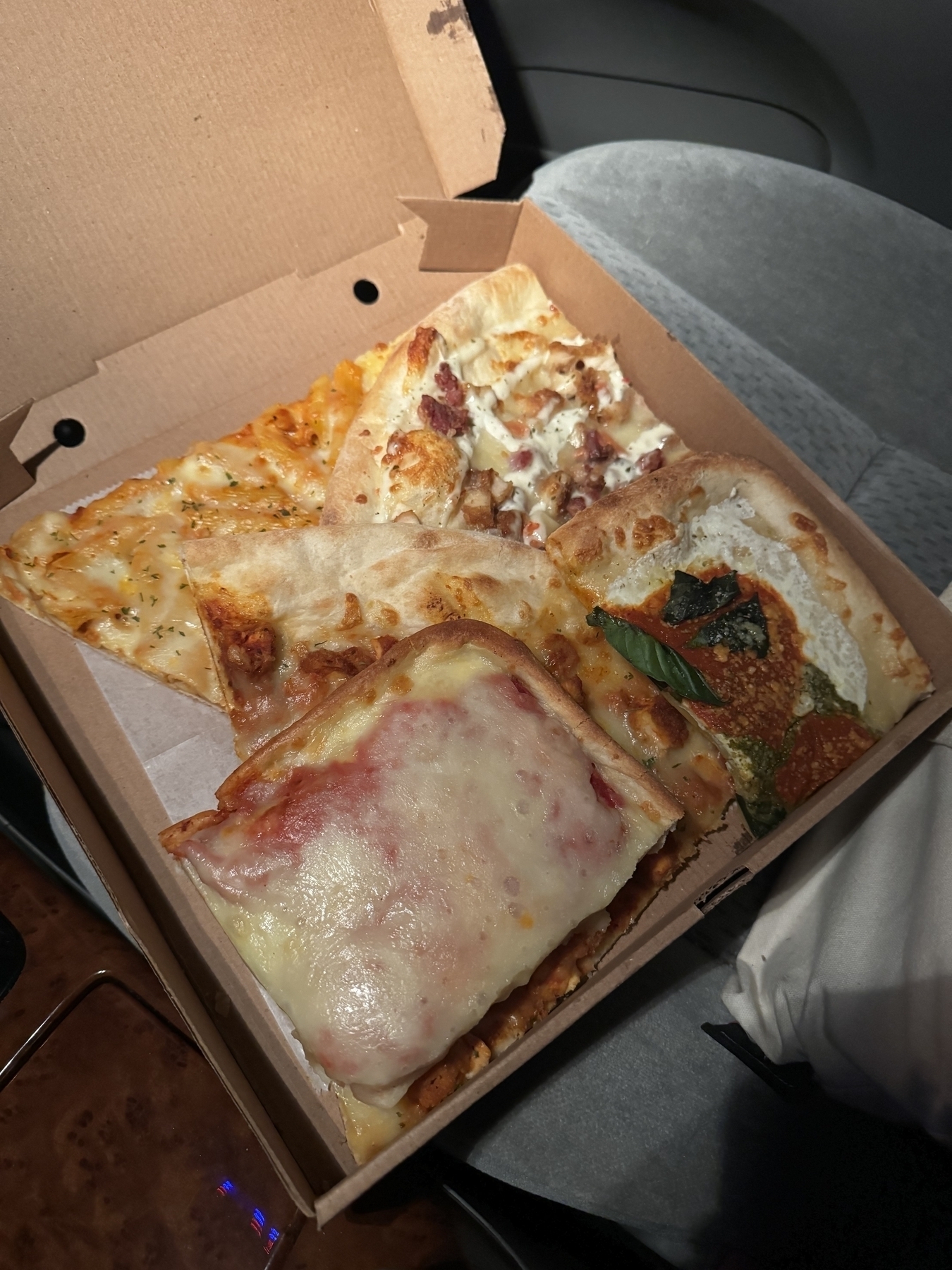 Five slices of pizza overlapping inside a square pizza box. Sat on a grey fabric car set. Illuminated by the ceiling light.
