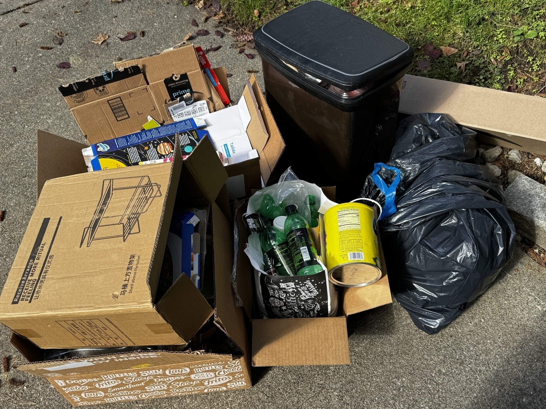Piles of recycling and a garbage bag and cardboard boxes filled with containers outside in the day time on concrete. 