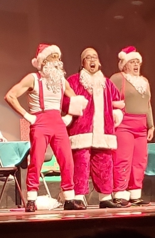 Three people dressed in Santa Claus costumes with sleeveless tops and red pants on a stage. They wear Santa hats and beards, and the central figure is singing or speaking with an open mouth, while the others appear to be in mid-performance.
