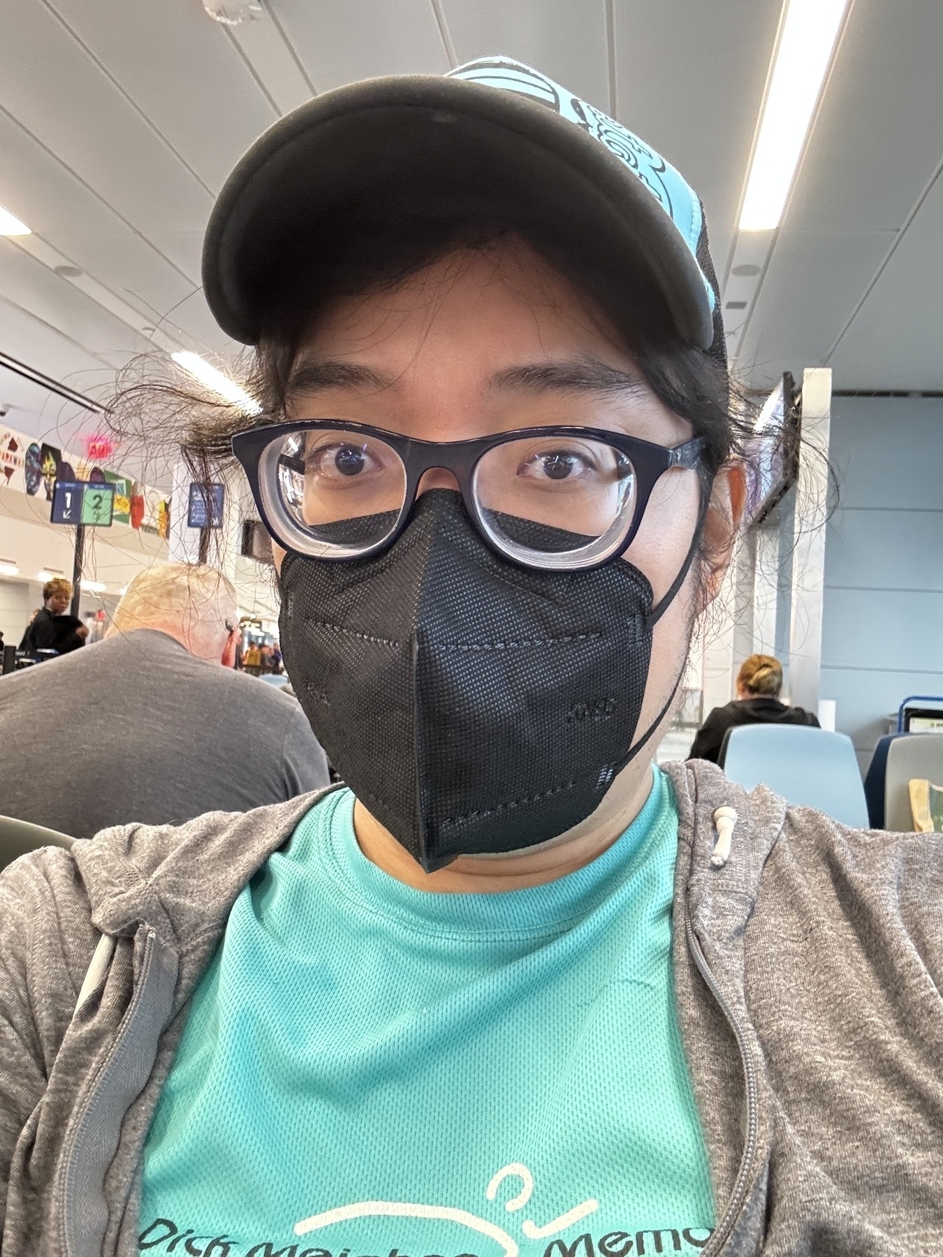 Miguel a Masc Filipino man with a real shirt and a teal hat in a black face mask in an airport gate 