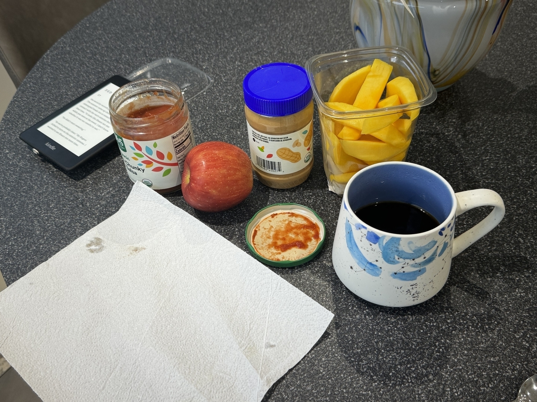 Food and a paper towel on a table. There’s an e reader. Salsa apple peanut butter mangos coffee black 