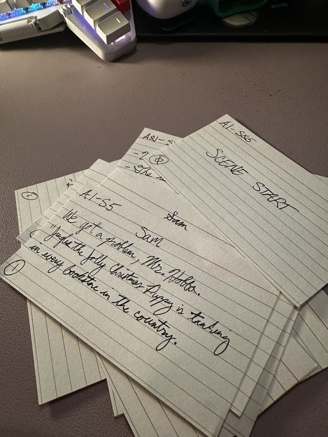 Index cards with handwritten script lines written on them. On a desk mat that is light purple. Part of a keyboard is in the background. 
