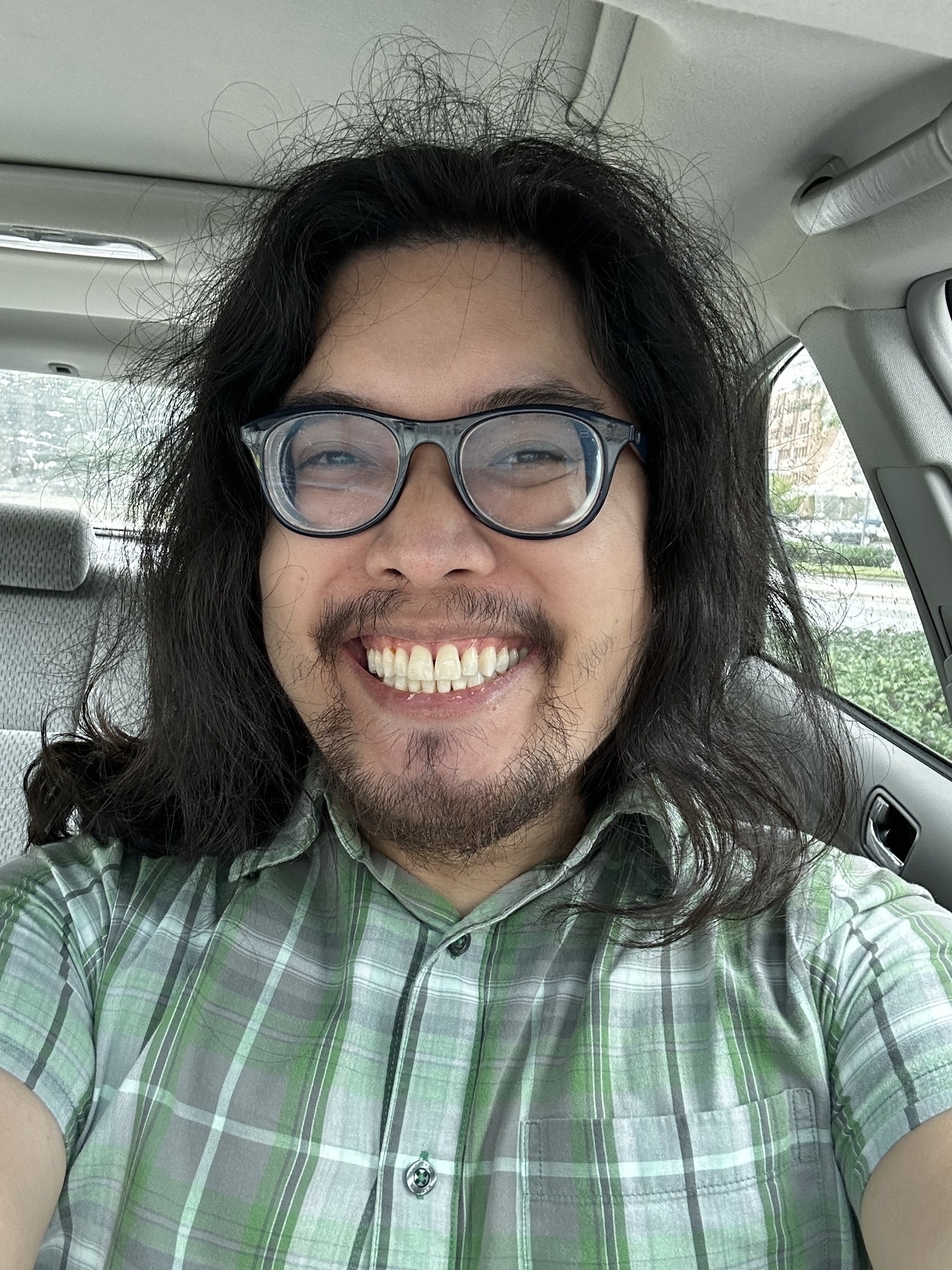 selfie of miguel a filipino man. he is smiling. he is inside a car. he has pong wavy black hair. he is wearing dark framed glasses. he is wearing a green shirt with buttons and a square pattern.