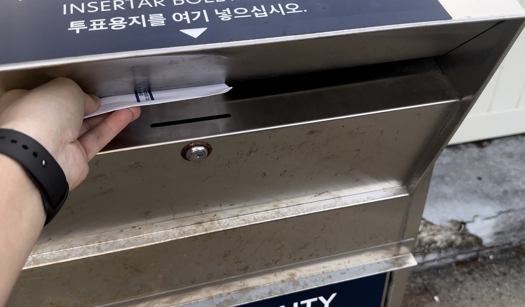 A man wearing a watch on his left arm is inserting an envelope into a shiny silver metal election drop box outdoors.  The envelope is most of the way in the slot. 