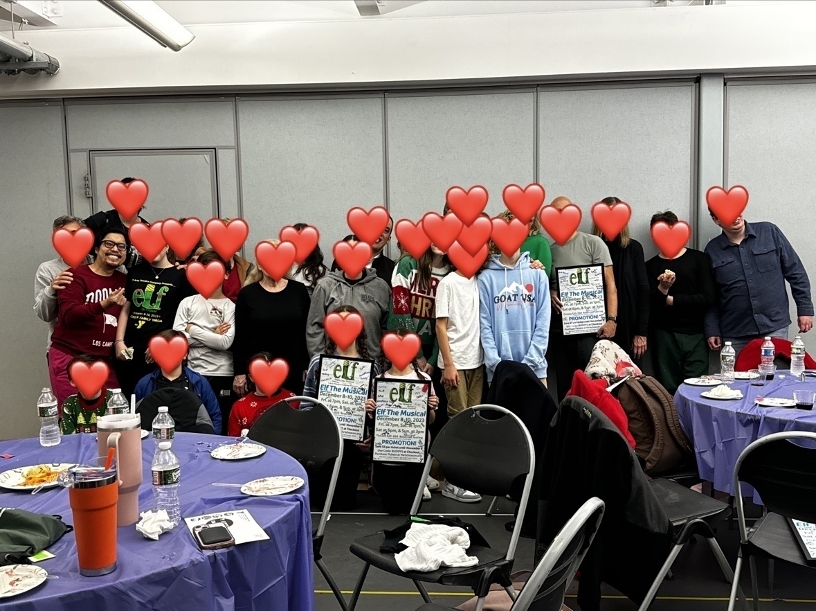 Group of people posing for a photo in a room with tables and chairs, faces covered by heart emojis.