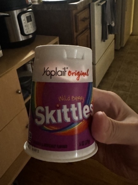 a right hand holding a contain of yoplait yogurt that is wild berry skittles flavour. it is indoors in a kitchen. an instant pot and stove are in the background.
