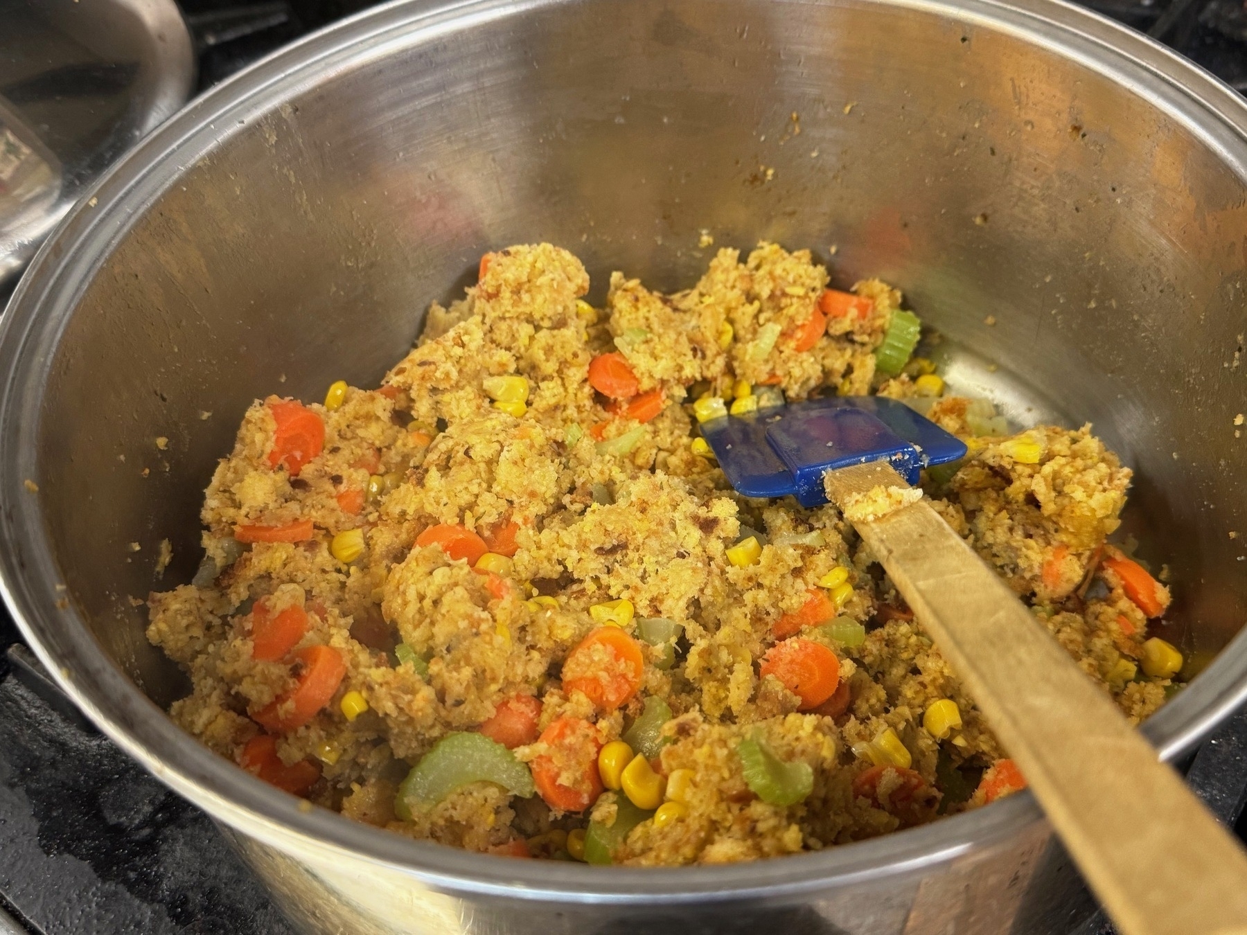 close up of stuffing from a box with carrots, celery, and corn in a silver steel pot with a wooden spatula sticking out of it