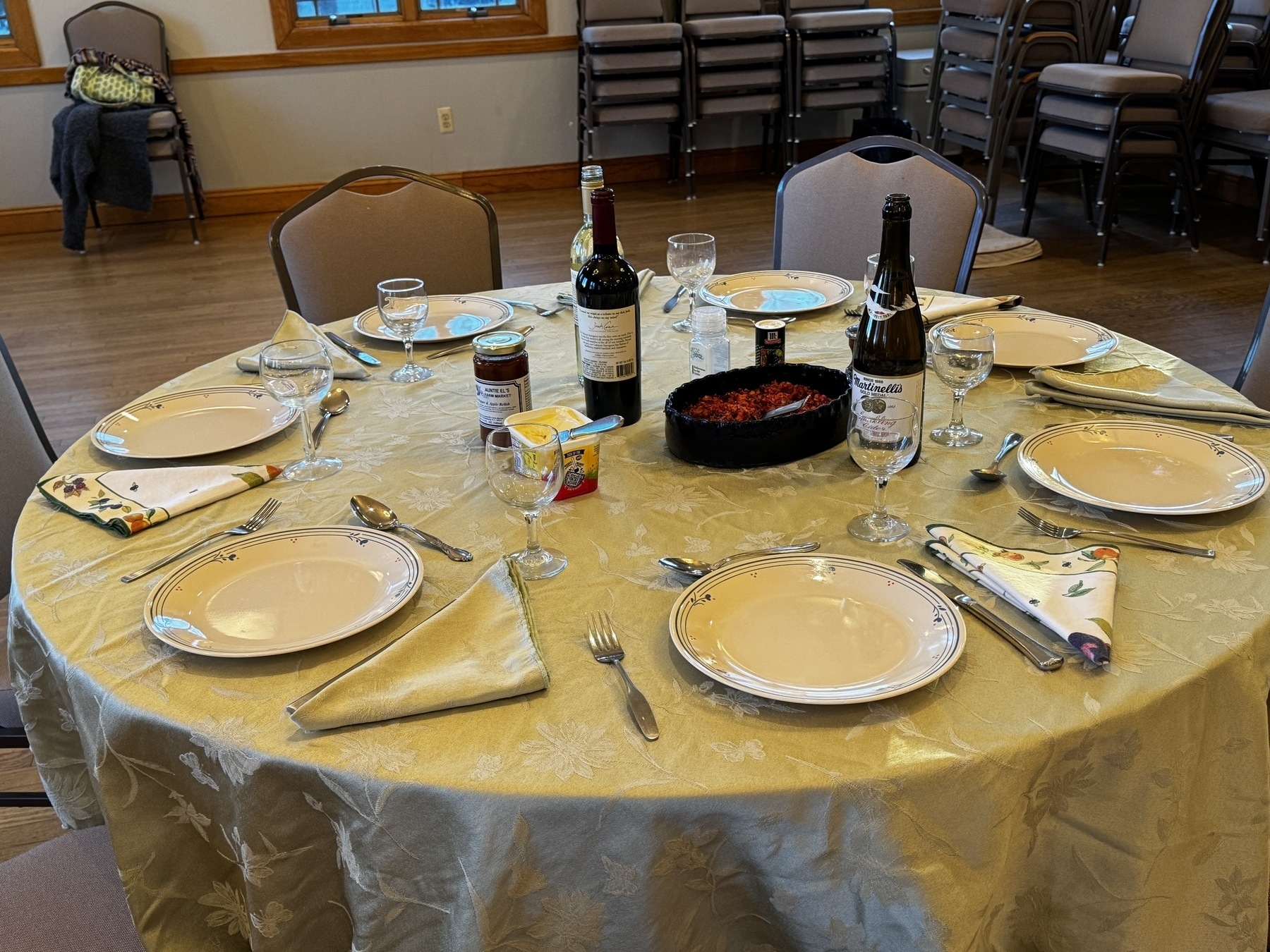 a table with empty chairs around it. a white table cloth is on a circle table. each place is set. there's glasses bottles and butter and a relish in the center.