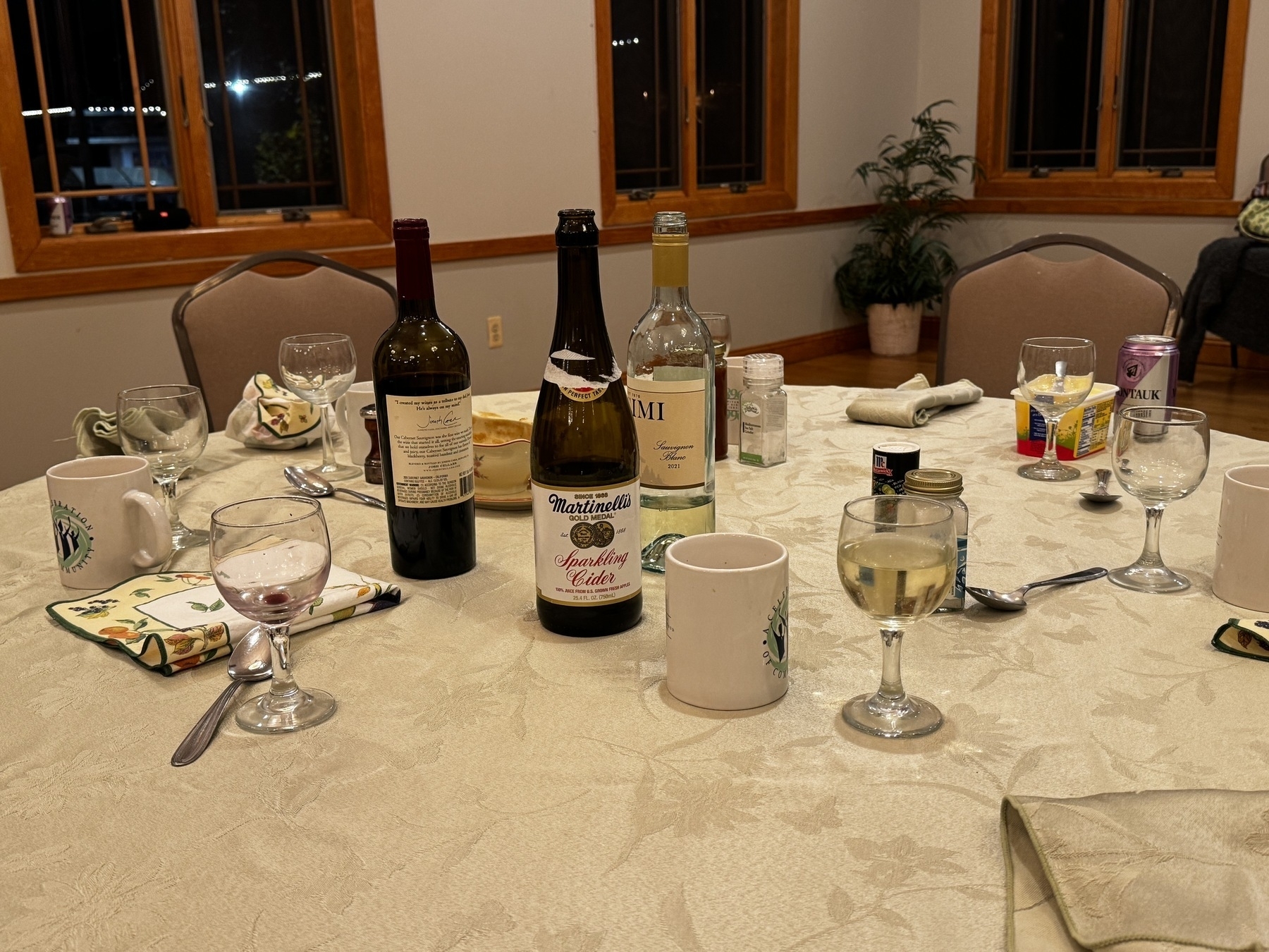 a set table at nighttime. there are three tall glass bottles. there are glasses and mugs. a white tablecloth. no one is sat down yet.