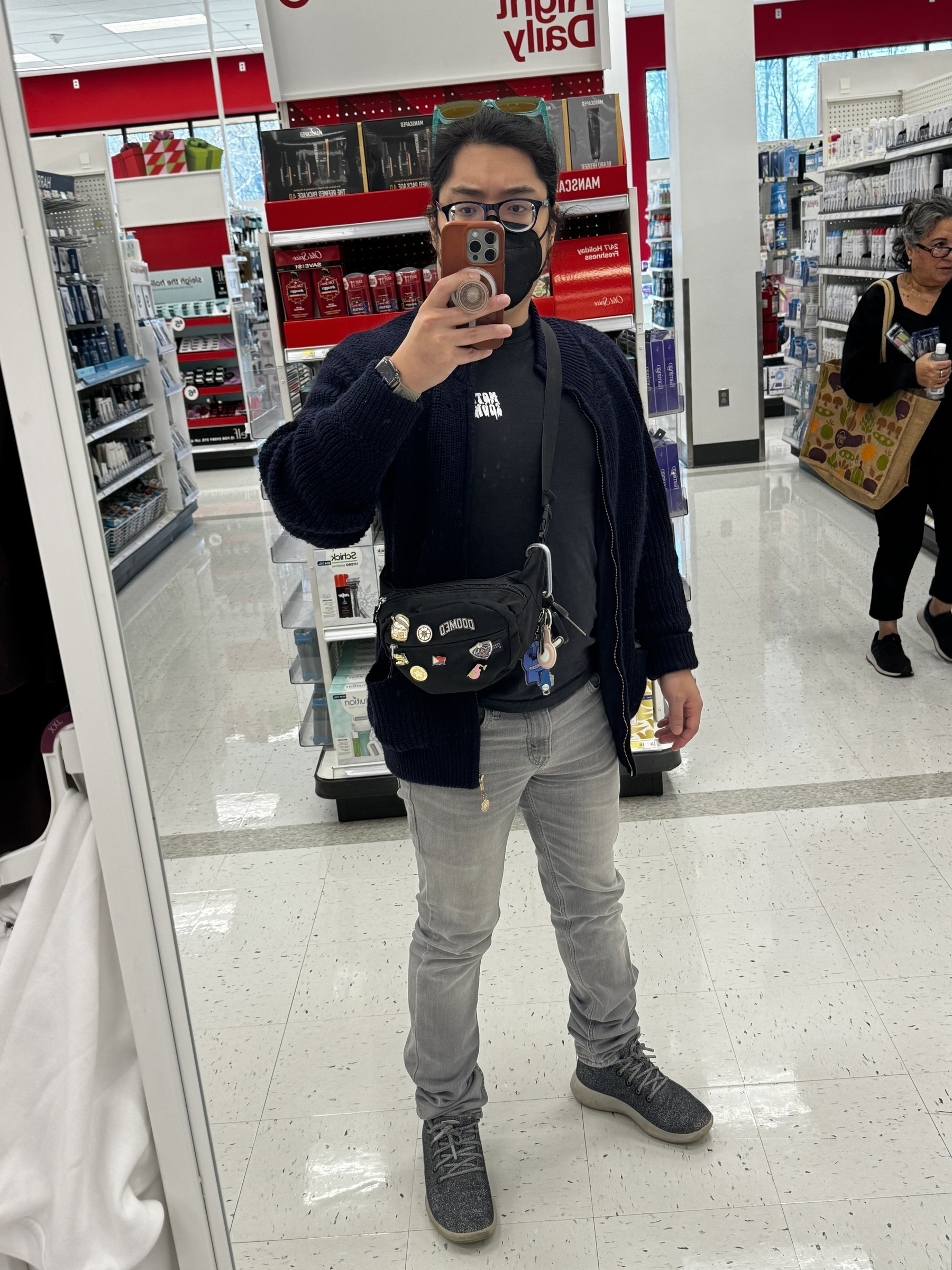Miguel a masc filipino man with black hair in a ponytail. Selfie inside a target in the daytime. wearing navy sweater, black shirt, black sling bag, grey jeans, grey shoes, black glasses, black mask.