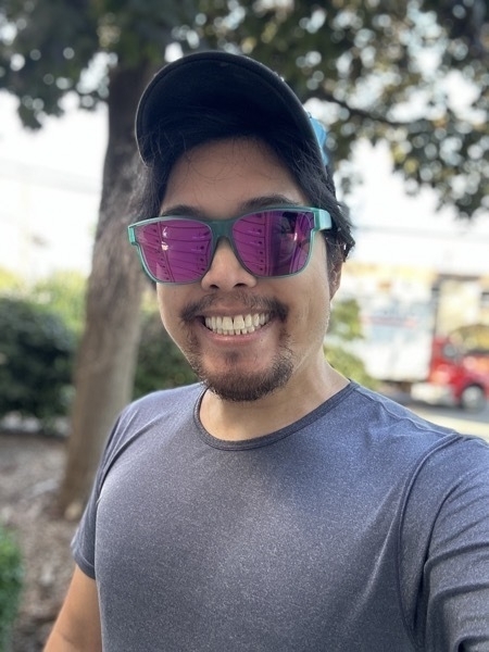 Miguel a masc filipino man is standing outdoors and smiling at the camera. he is wearing a blue short sleeve shirt. it is daytime. he is wearing a trucker hat. the sunglasses are mint green frames and purple lenses. miguel has a mustache and a goatee. he has great teeth.
