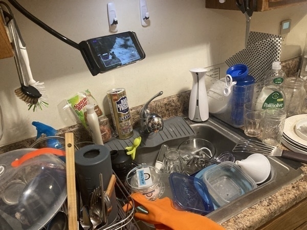 a sink full of dishes. to the left is a dish drying rack that's full. a phone is suspended on a bendable arm for viewing.