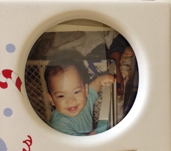 baby photo of Miguel a filipino boy. he is under two years old probably. he is smiling. he is wearing a blue shirt. he is in a play pen. he has hair. the frame the photo is in is Christmas themed.