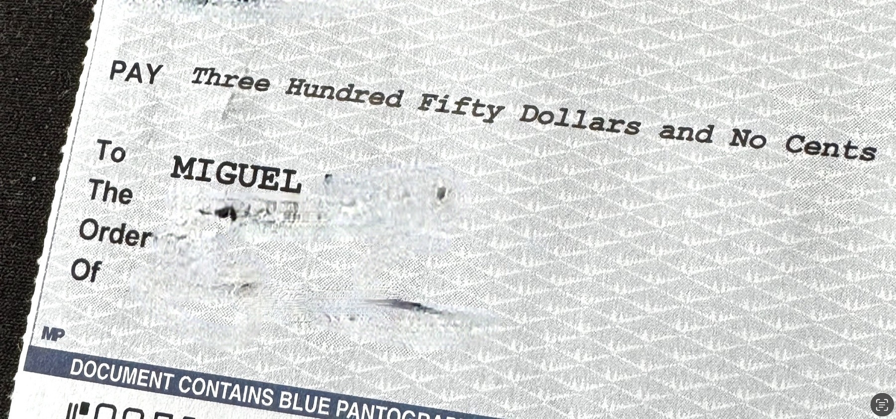 Close-up of a check with the text "Pay Three Hundred Fifty Dollars and No Cents" and "To the Order of MIGUEL". The check features a security pattern and text stating "DOCUMENT CONTAINS BLUE PANTOGRAPH".