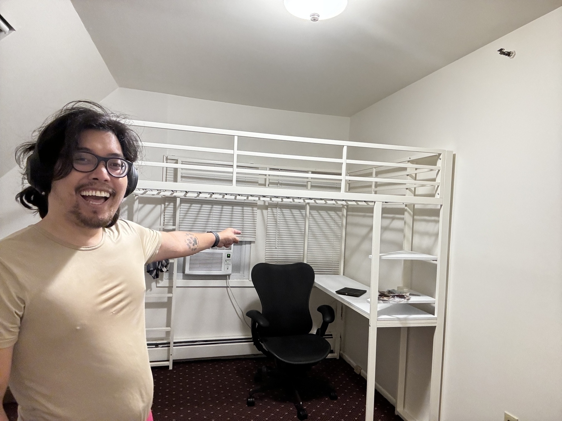 A person with glasses and headphones, smiling and pointing at a loft bed with an integrated desk and shelves in a small room. The room has white walls, an air conditioner, and a dark carpet.