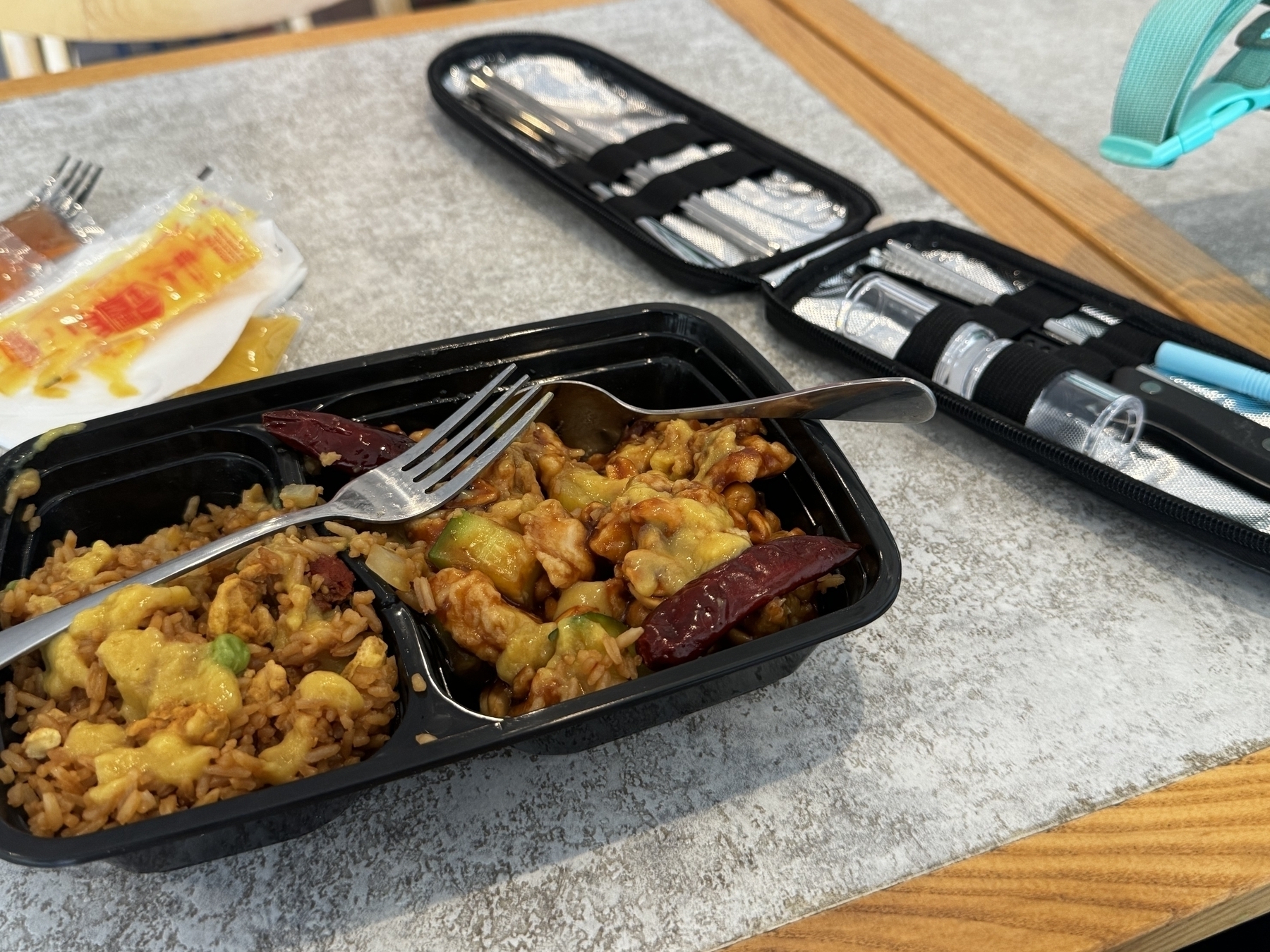 A meal consisting of fried rice and a stir-fry dish with vegetables and chicken in a black container, accompanied by metal utensils and a set of chopsticks, spice shakers, and a straw in a black case.