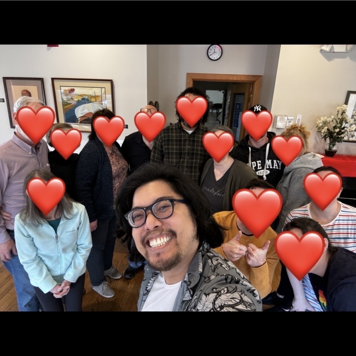  A group of people of all ages are posing for a photo in a room. The people are all smiling and some are making peace signs. There are red heart emojis covering the faces of the people in the photo.