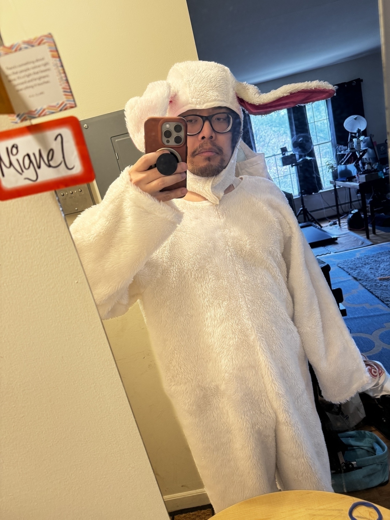  A person wearing a white bunny costume with a pink inner ear and black glasses is taking a selfie in front of a mirror.