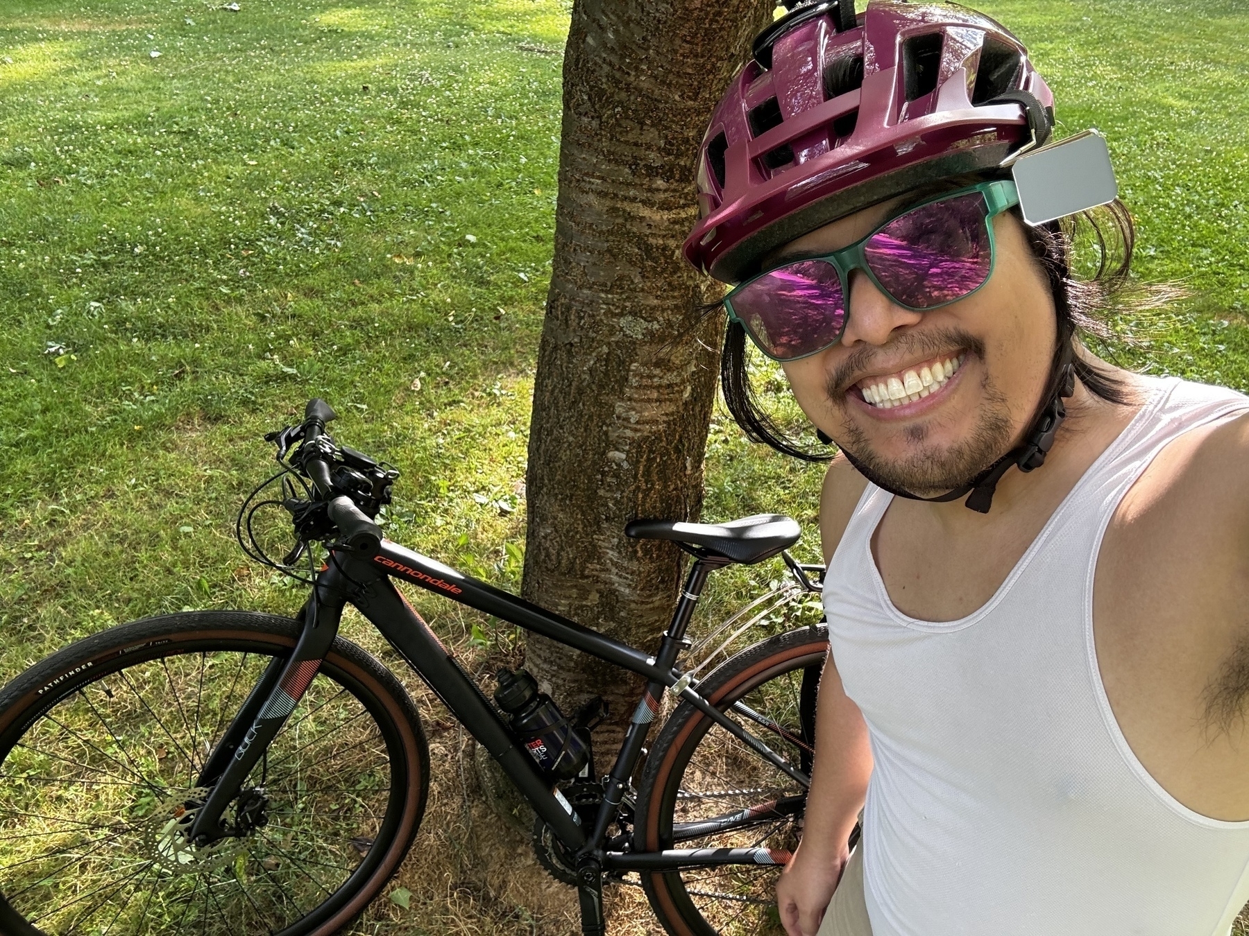 A person wearing a pink bike helmet and sunglasses is smiling while taking a selfie next to their black Cannondale bike, which is leaning against a tree. The person is dressed in a white tank top, and the background features a grassy park area.