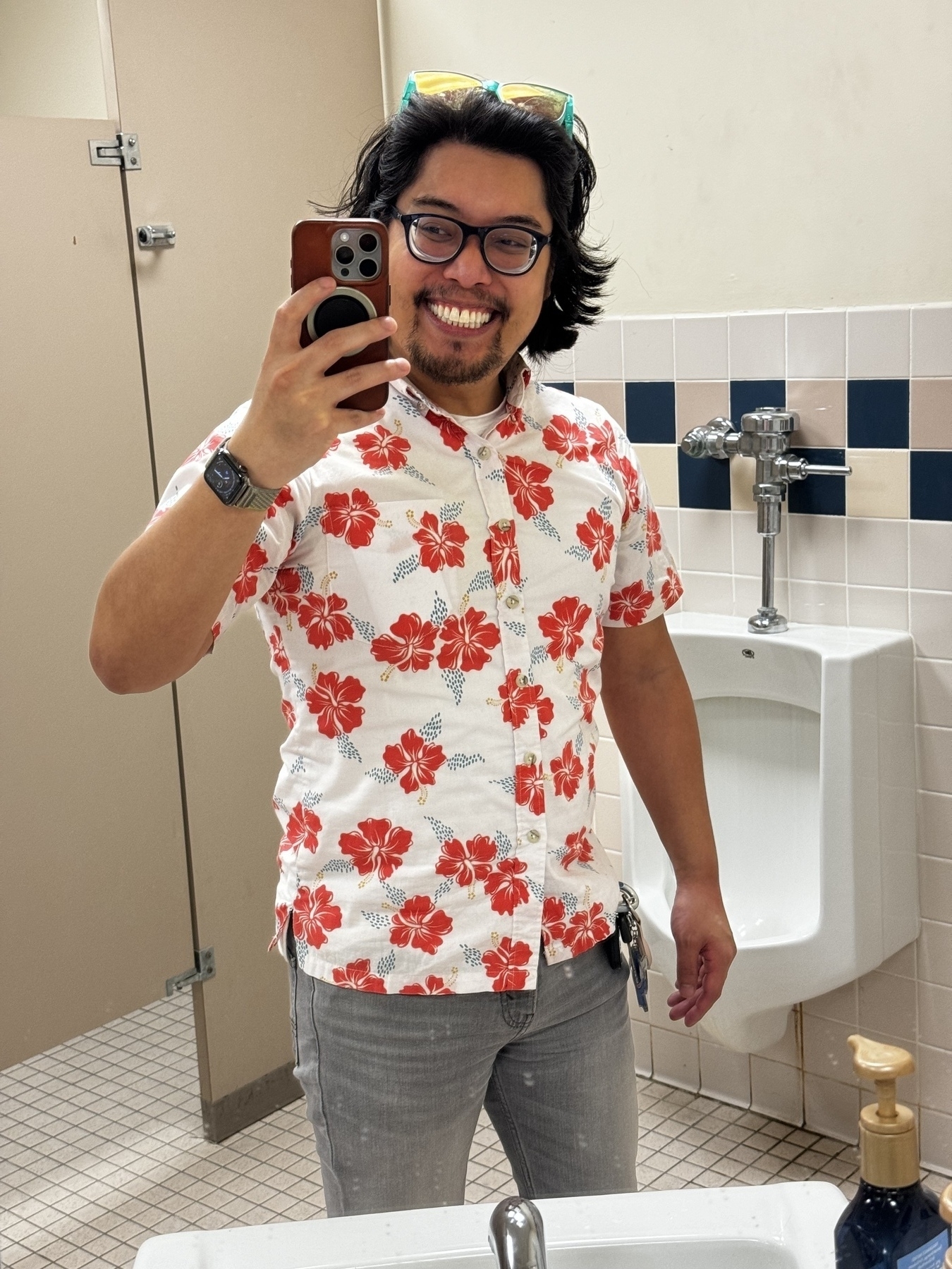A smiling man wearing glasses, a floral shirt, and jeans takes a selfie in a bathroom.