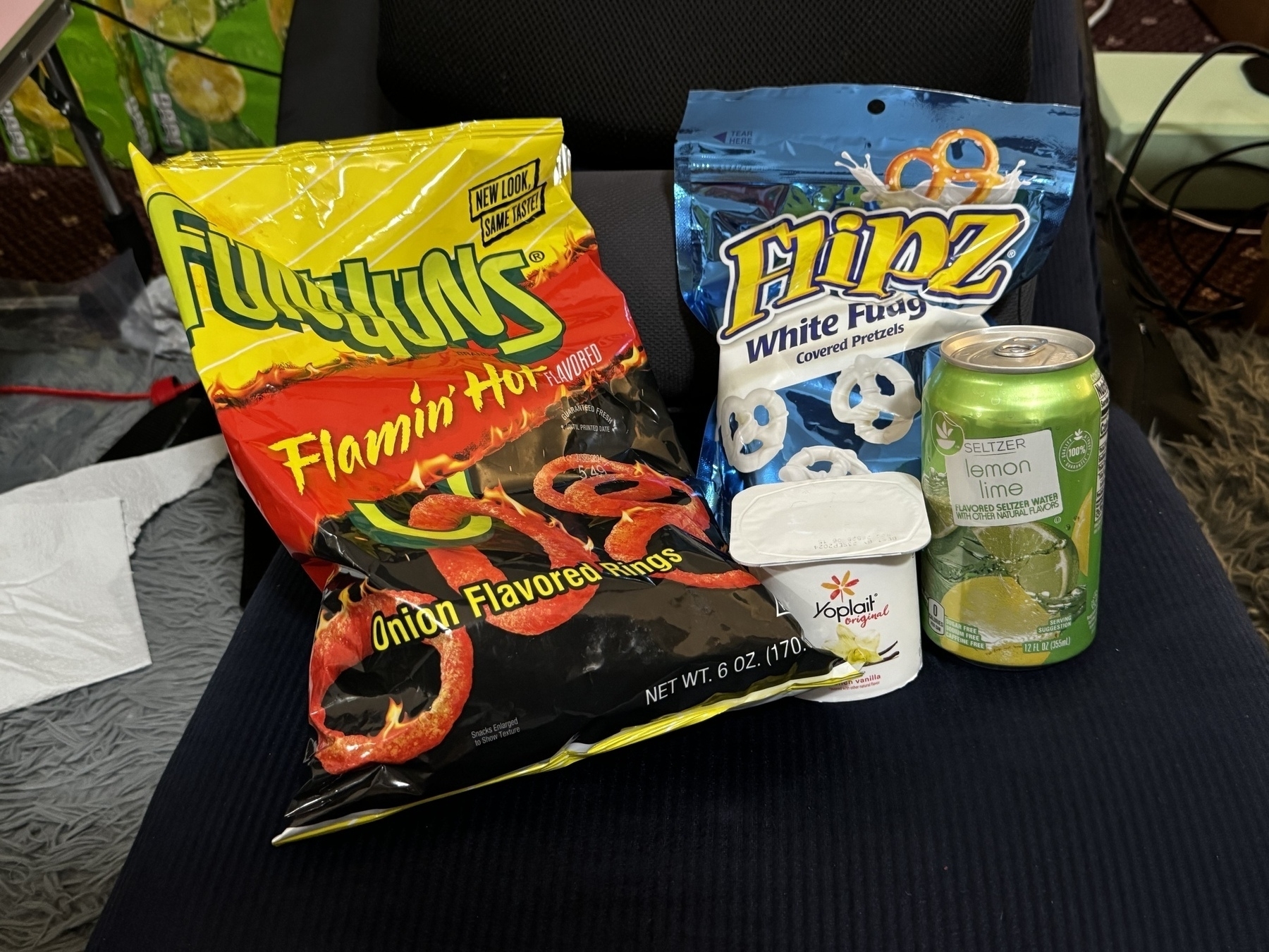 The image shows a selection of snacks arranged on a surface. The items include a bag of Flamin' Hot Funyuns (onion-flavored rings), a bag of Flipz White Fudge-covered pretzels, a container of Yoplait Original vanilla yogurt, and a can of lemon-lime flavored seltzer water. The snacks are placed on a dark chair or cushion, with additional items like a tissue and boxes partially visible in the background.