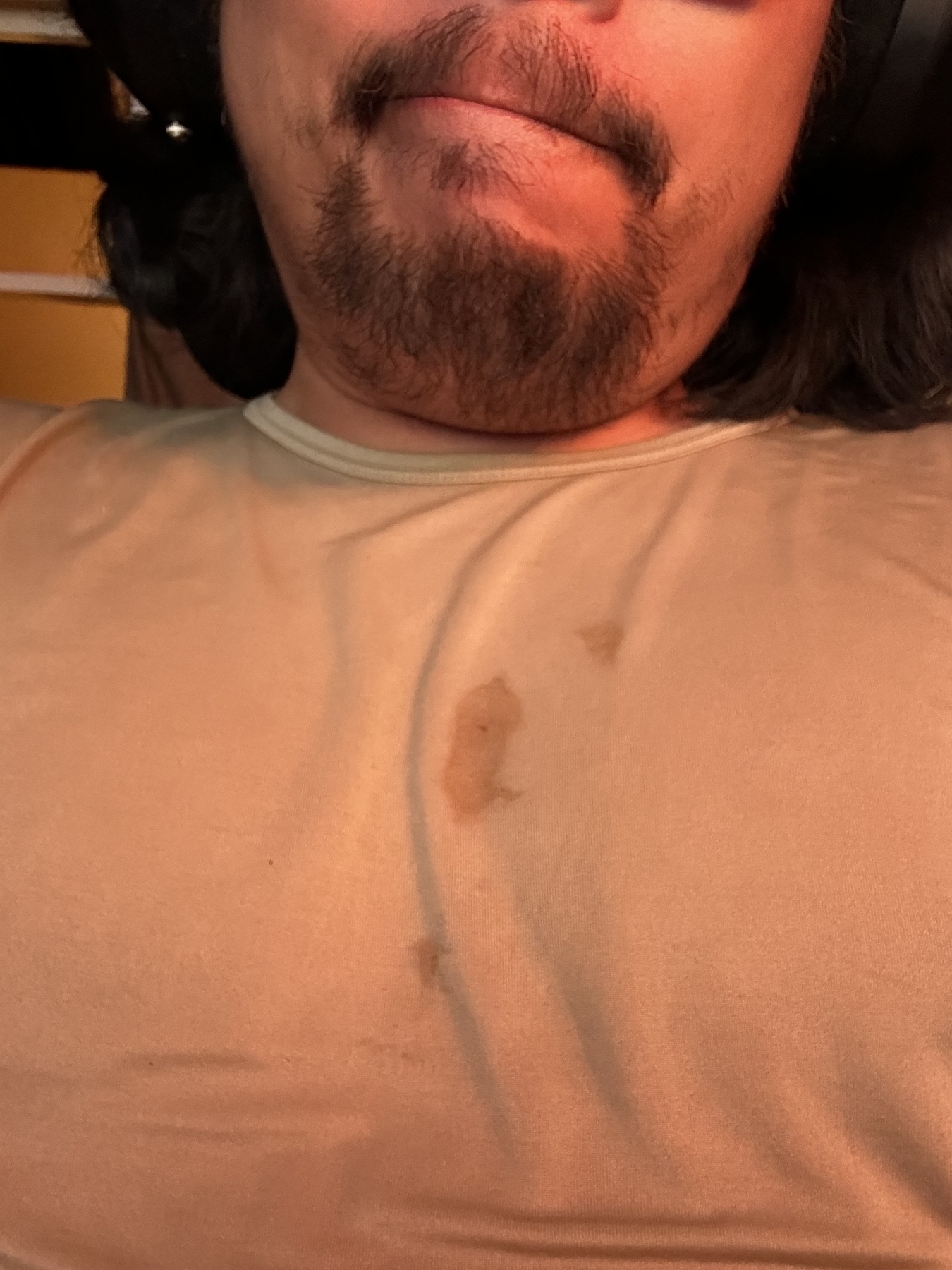 The image shows a close-up of a person's lower face and upper chest. The person has a beard and mustache, and is wearing headphones. They are wearing a beige or light-colored shirt that has visible stains on the chest area. The stains appear to be from some type of liquid or food.