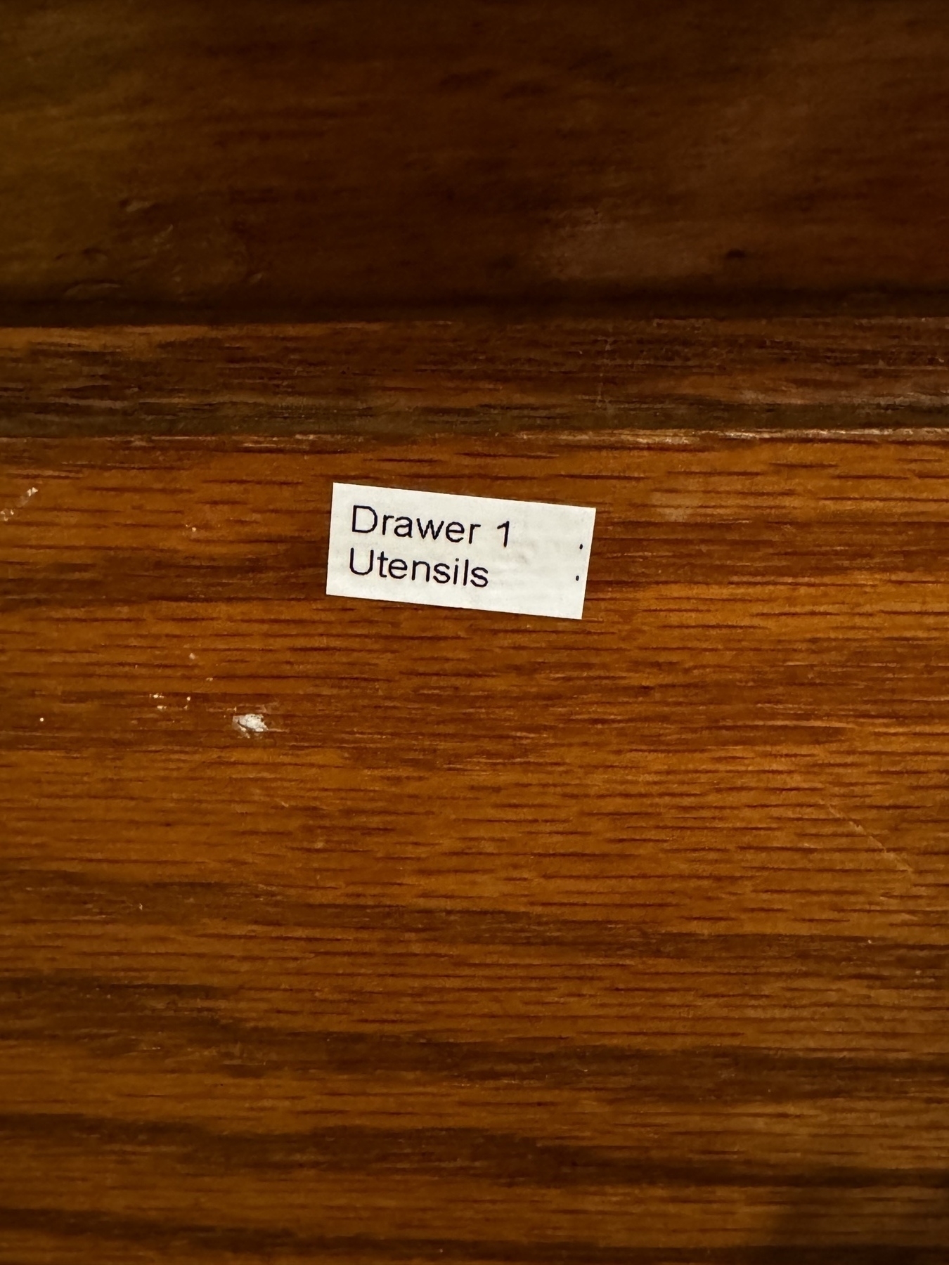A small white label with black text reading "Drawer 1 Utensils" is adhered to a woodgrain surface.