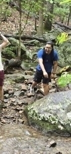 A person hiking through a forested area, wearing a blue shirt and black shorts, smiling and navigating over rocks and a small stream, with another person partially visible on the left side.