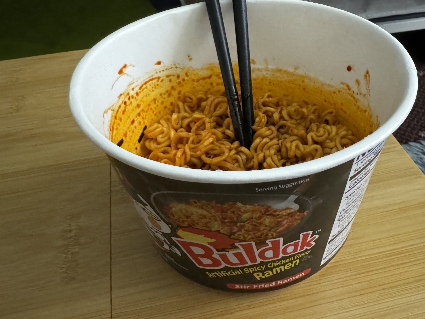 The image shows a cup of instant noodles labeled "Buldak Artificial Spicy Chicken Flavor Ramen" with chopsticks resting inside. The noodles are coated in a deep red sauce, suggesting a spicy flavor. The cup is sitting on a wooden surface.