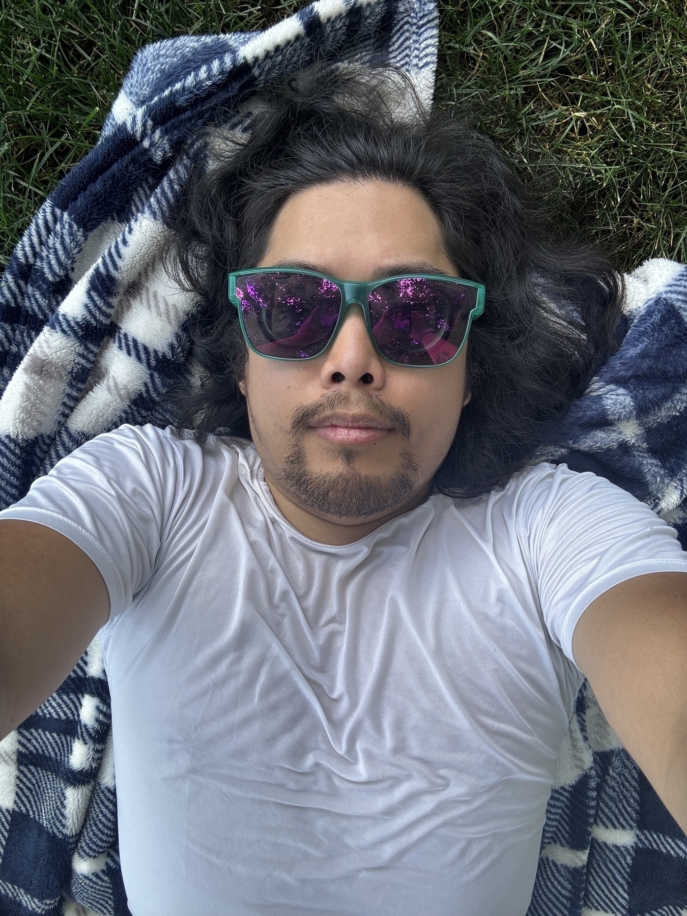 A person lying on a checkered blanket in the grass, wearing a white shirt and green sunglasses with reflective lenses. They have shoulder-length hair and a short beard.