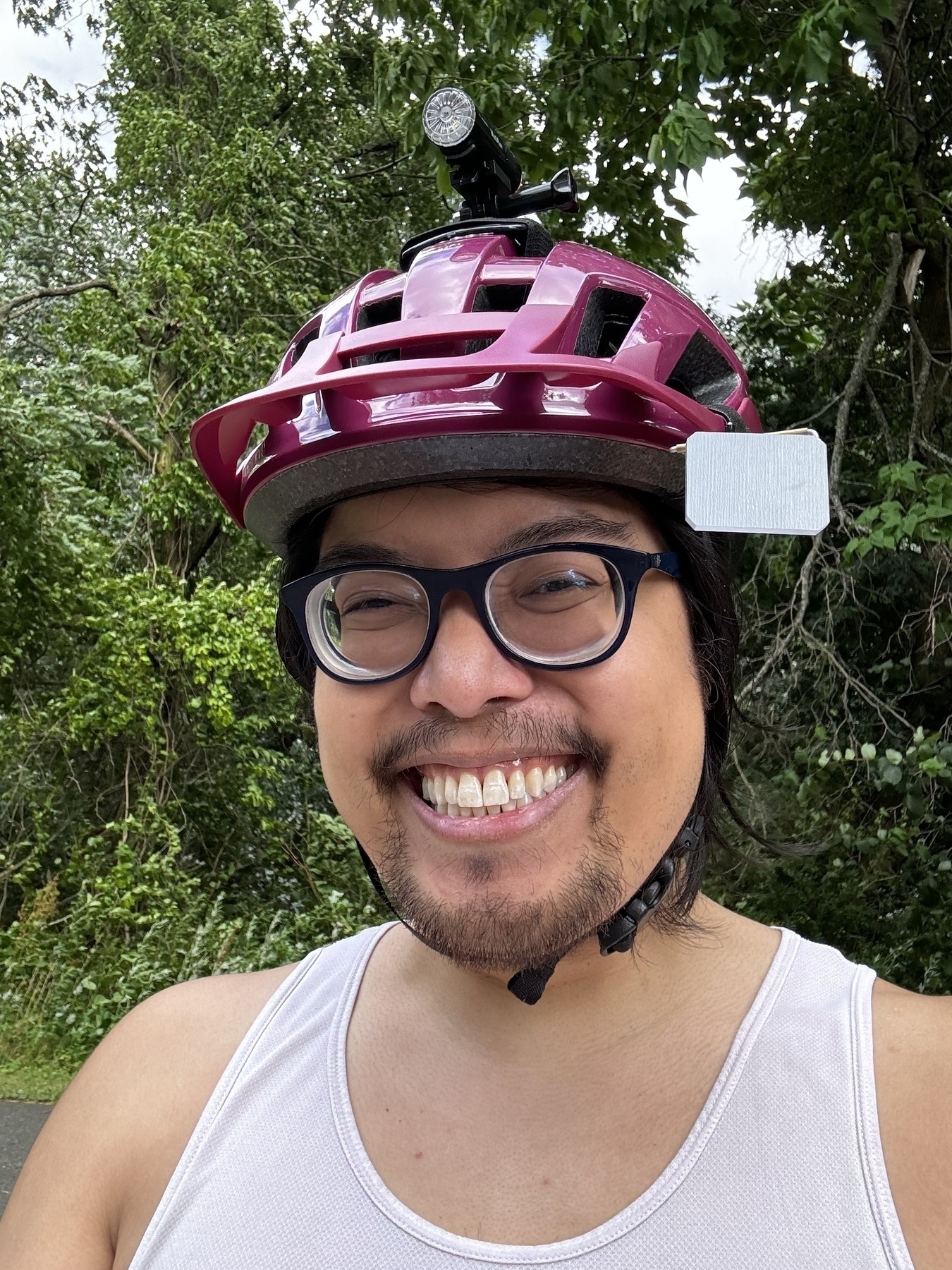 The image shows a Filipino man wearing a bright pink bike helmet with a mounted light on top, a white tank top, and glasses, smiling broadly while outdoors. The background features lush green foliage, indicating they are likely on a bike ride in a natural setting.