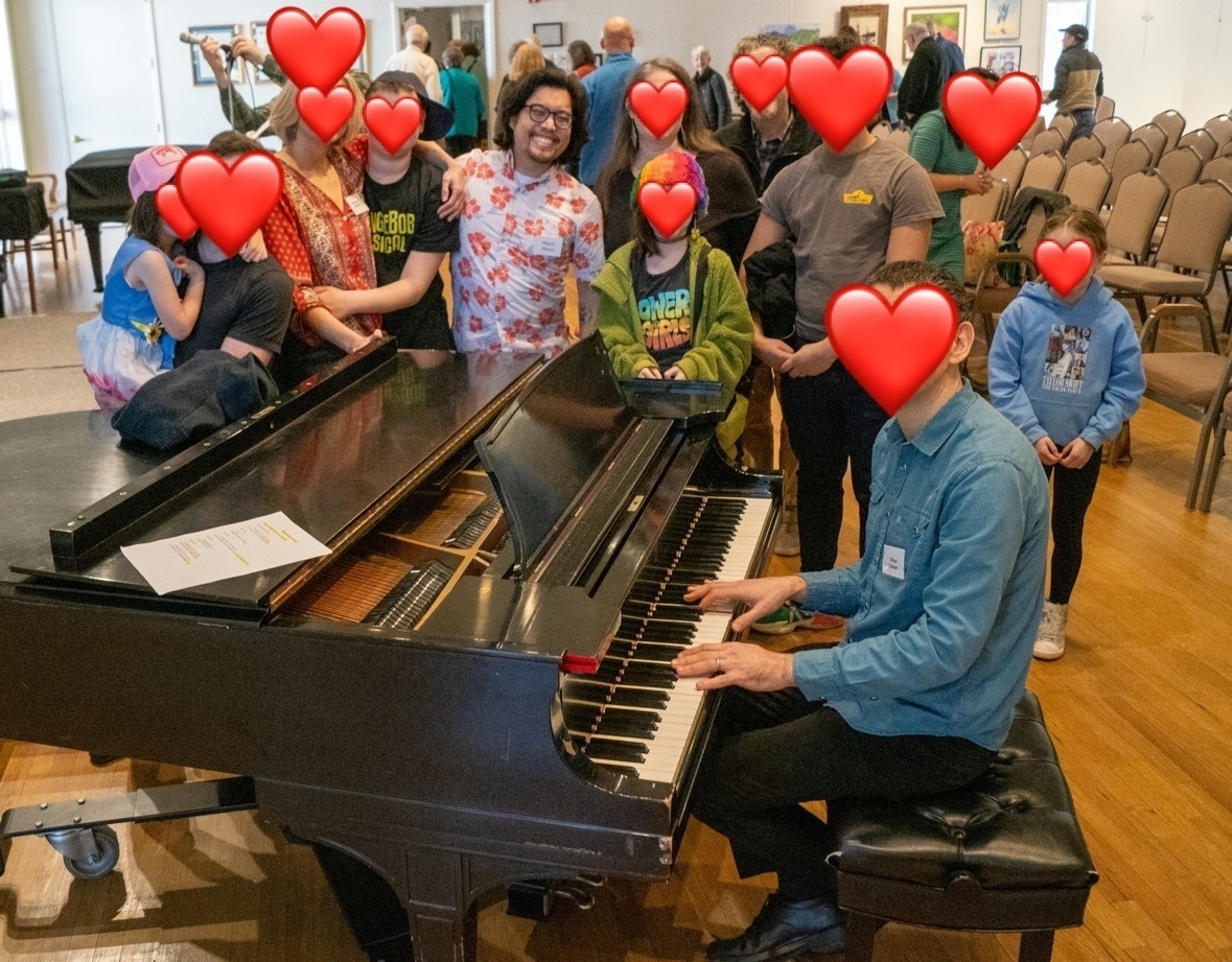 A group of people are gathered around a piano in a room. Some of the people are sitting on chairs, some are standing. A man is sitting at the piano