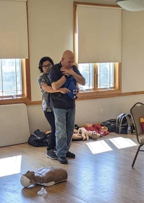 Two men are standing. One is practicing giving the heimlich maneuver. They are indoors and it is daytime. There is a CPR dummy on the floor.