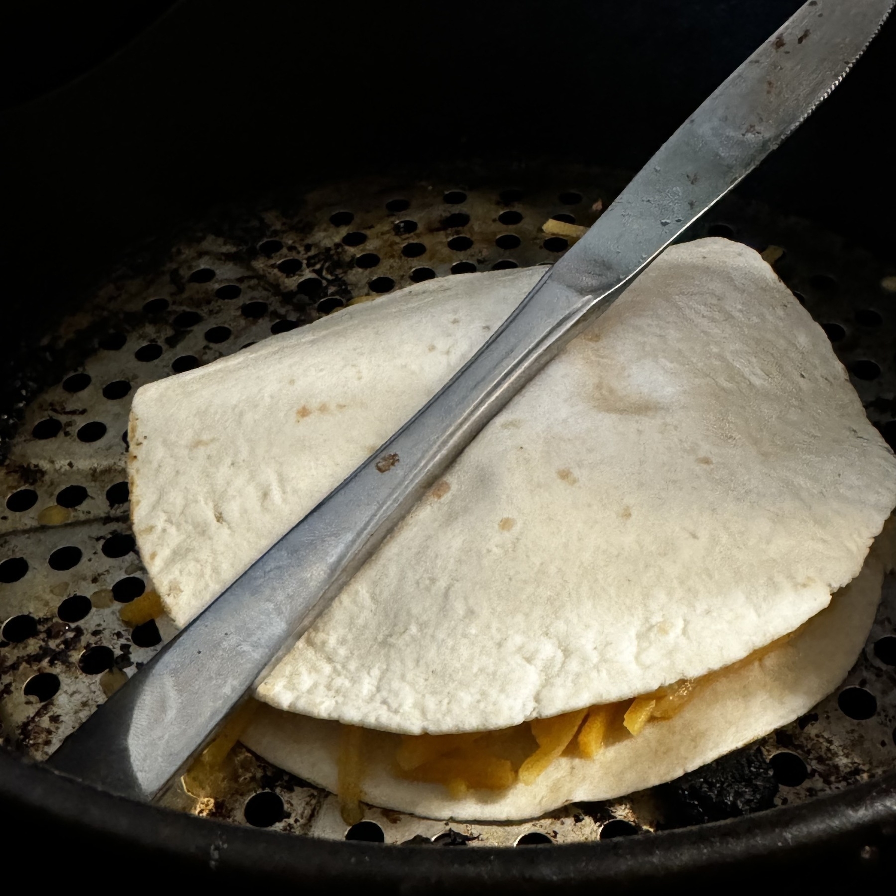 A knife is on top of a quesadilla in an air fryer basket. The quesadilla is made with two tortillas and cheese and sauerkraut