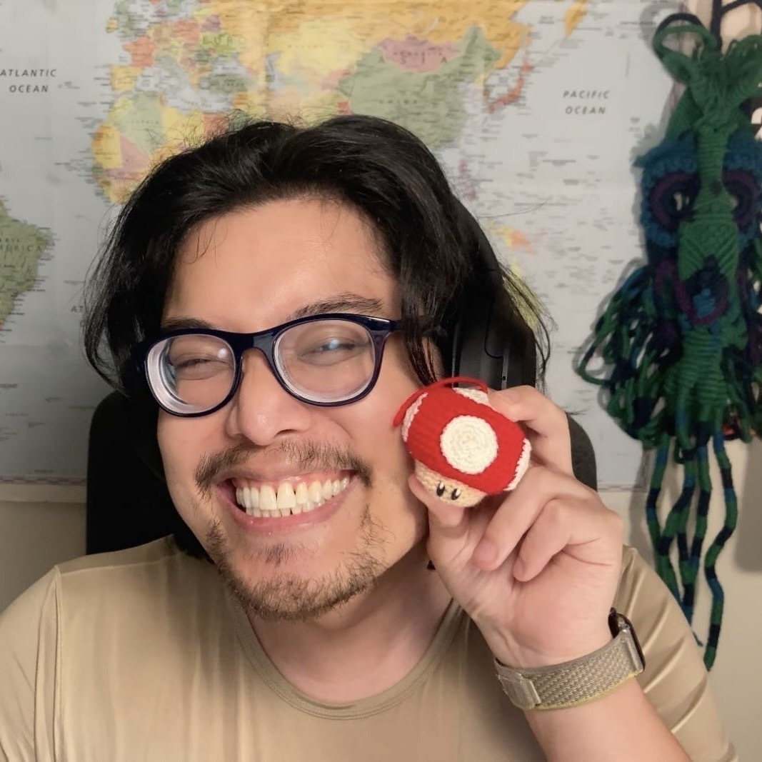 A young man with brown hair and glasses is smiling and holding a small, red and white mushroom toy.