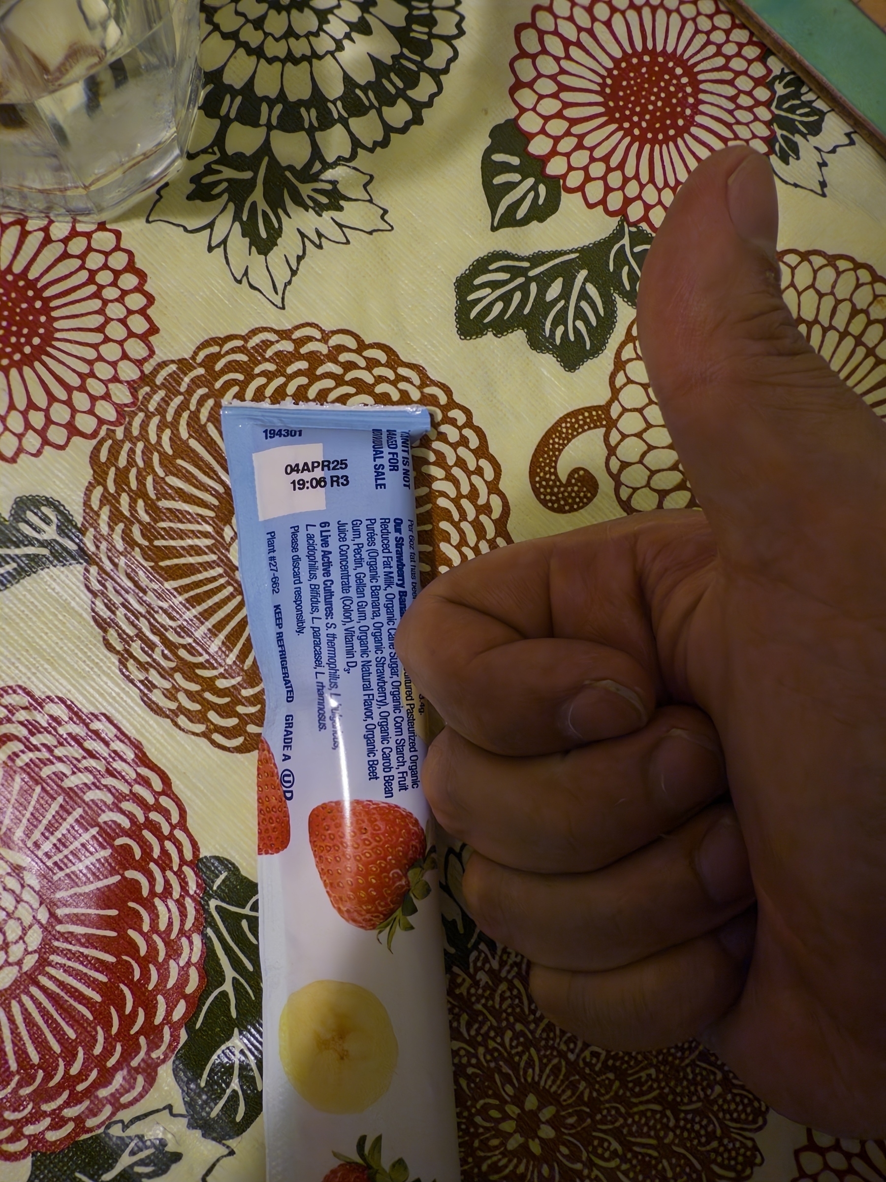 Hand makes a thumbs up next to a white yogurt package with strawberry and banana images against vibrant floral tablecloth. Colorful dahlia patterns in red, brown, and green create bold backdrop. Glass and napkin visible in corner.​​​​​​​​​​​​​​​​ expiration April 2025