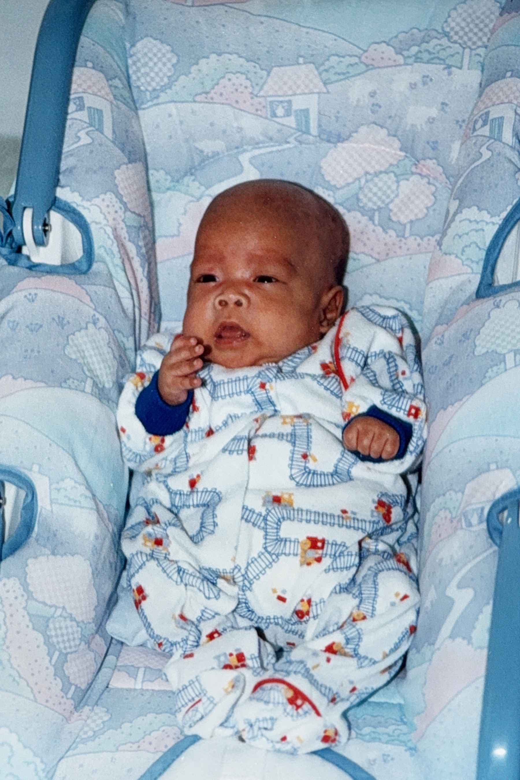 Newborn baby lies in blue car seat, wearing white onesie with colorful train pattern, left hand raised to mouth. Soft pastel village scene with houses and clouds decorates the seat's fabric background, creating a gentle, dreamy atmosphere.​​​​​​​​​​​​​​​​